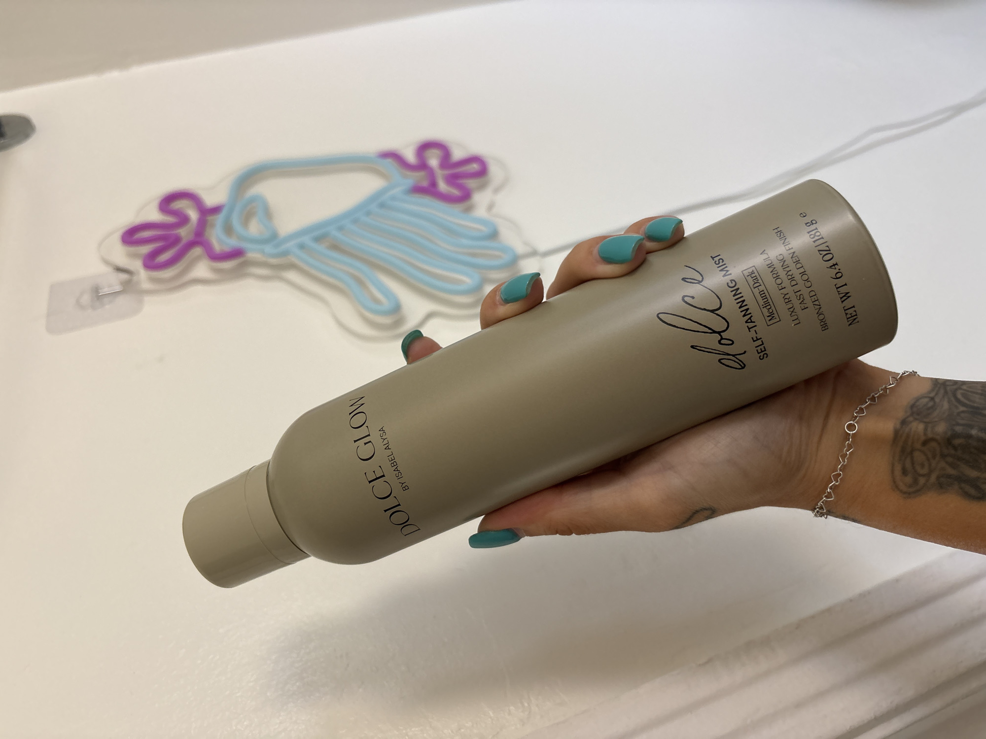A close-up, indoor shot from Seal Beach, USA, features a person's hand holding a cylindrical bottle of self-tanning mist against a plain white background. The hand, adorned with teal nail polish, a delicate silver heart-link bracelet, and a visible tattoo on the forearm, grips the bottle.
The bottle, in a matte, light beige or taupe color, is angled across the frame. Prominent text on the bottle reads "DOLCE GLOW BY ISABEL ALYSA" vertically on one side, and "dolce" in a stylized script, followed by "SELF-TANNING MIST". Further details on the bottle specify "Medium-Dark", "LUXURY FORMULA", "FAST DRYING", and "BRONZED GOLDEN FINISH". The net weight is stated as "NET WT 6.4 OZ / 181 g e".
In the soft-focus background, a decorative neon sign shaped like a cloud with raindrops is visible. The cloud outline is purple, and the raindrops are light blue, mounted on a clear acrylic base. The overall setting appears to be a clean, bright indoor space, possibly for a product showcase or personal use.