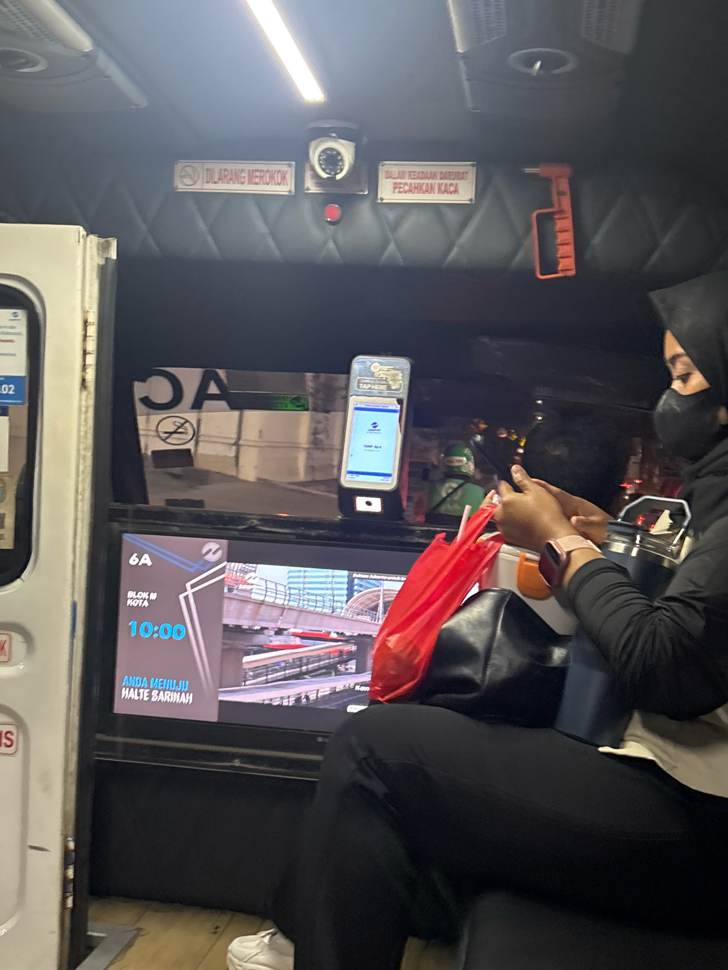 A low-angle, eye-level shot captures the interior of a public transportation vehicle, likely a bus or commuter train, as suggested by the seating and overhead fixtures. The scene is dimly lit, illuminated by a bright overhead fluorescent light and the glow from a digital display.
In the foreground, a person wearing a black hijab and a black face mask is seated on the right, facing left. They are dressed in black clothing and have a pink smartwatch on their left wrist. Their hands are engaged with a smartphone, which is held between them. A bright red plastic bag sits on their lap, and a dark blue tumbler is to their right.
On the left side of the image, a white panel with some text and numbers is visible, along with a section of a screen displaying information about a route: "6A BLOK M KOTA 10:00 ANDA MENUJU HALTE SARINAH". This suggests the location is within North Jakarta, Indonesia, likely on a public transport route connecting to the Sarinah area.
Above the screen, a digital display shows an image of an elevated railway system. Further up on the ceiling of the vehicle, two signs are prominently displayed. The left sign reads "DILARANG MEROKOK" (No Smoking), with a crossed-out cigarette symbol. To its right, a circular camera is mounted, and below it, a red light is visible. The right sign reads "DALAM KEADAAN DARURAT PECAHKAN KACA" (In Case of Emergency, Break Glass), accompanied by an image of a hammer. An orange emergency hammer is mounted to the right of this sign.
The overall environment suggests a functional public transport interior, with a focus on safety and passenger information. The presence of the route information in Indonesian, combined with the "No Smoking" and emergency instructions, firmly places the scene in Indonesia, and the specific route details point to North Jakarta.