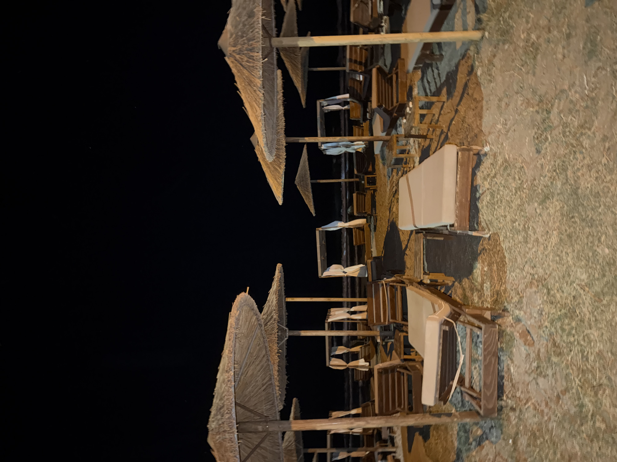 The image depicts a beach scene at night in Georgioupoli, Greece. The foreground is dominated by several empty wooden lounge chairs with beige cushions, some of which are positioned under thatched umbrellas. In the mid-ground, more lounge chairs and canopy-like structures with white drapes are visible. The sea is faintly discernible in the background, its surface reflecting some light from an unseen source. The sky is completely dark, with a few faint stars visible. The overall impression is of a quiet, deserted beach at night, likely a resort or public beach area.