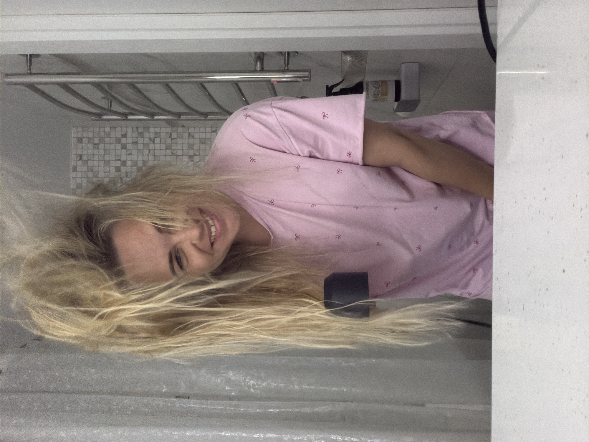 A woman with long blonde hair, wearing a pink shirt with small flower designs, is smiling at the camera. She appears to be in a bathroom. Behind her is a towel rack, a tiled wall, and a shower curtain. I cannot determine if this is Votkinsk, Russia, or anywhere else without additional information.