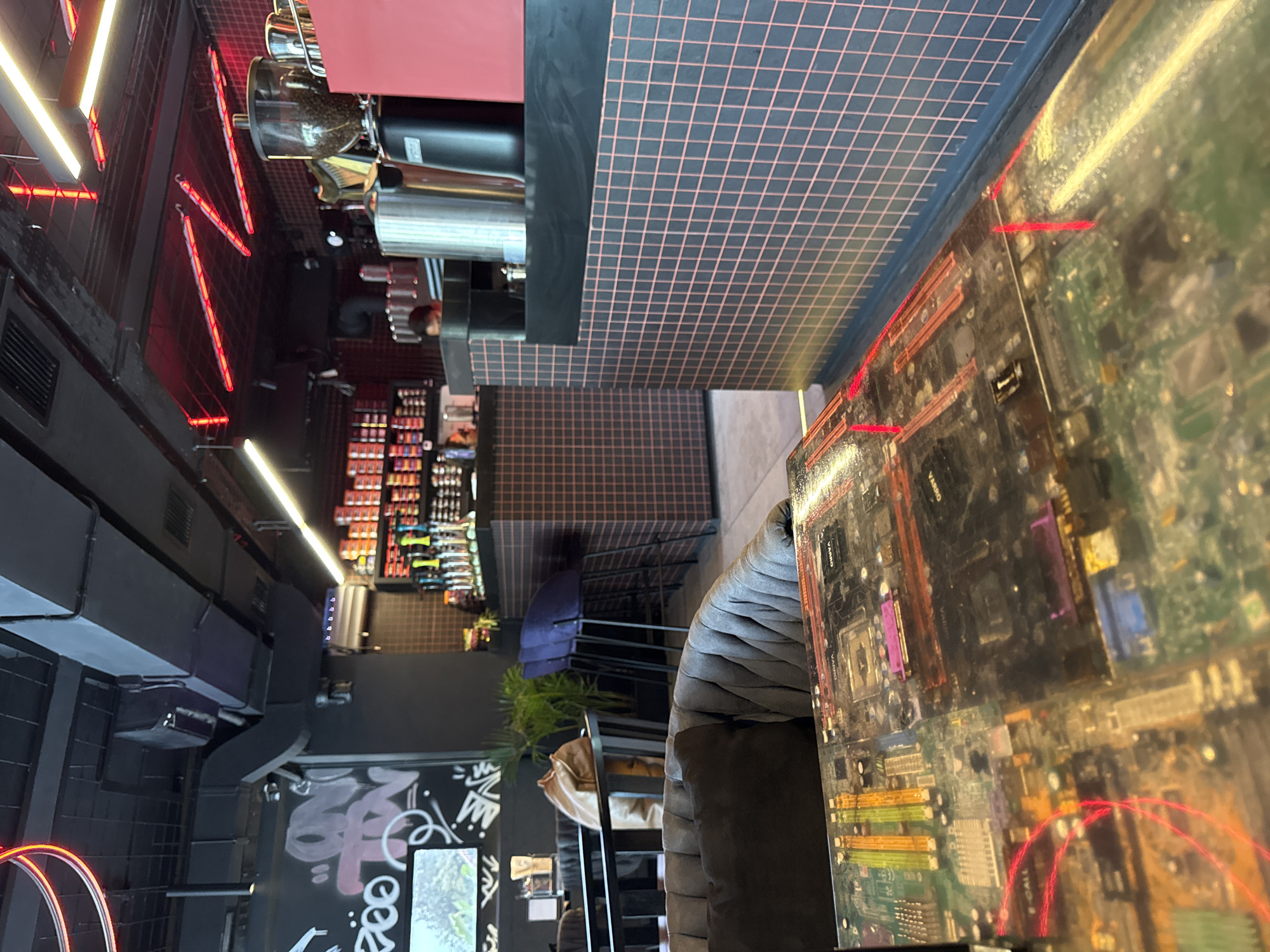 The image shows the interior of a cafe. The table in the foreground is covered with a transparent surface displaying a motherboard. Behind the table, a grey armchair with black and grey cushions is visible. Further back, a counter area is set up with a coffee machine and various bottles or containers. The walls are decorated with a grid pattern in pink and black. Red and white neon lights are present on the ceiling, along with ventilation ducts. There is a shelf displaying rows of colorful items, possibly coffee pods or tea boxes. Graffiti art is visible on a black wall in the background. A person is partially visible behind the counter. The overall aesthetic is modern and industrial, with a touch of vibrant color from the lighting and decor.