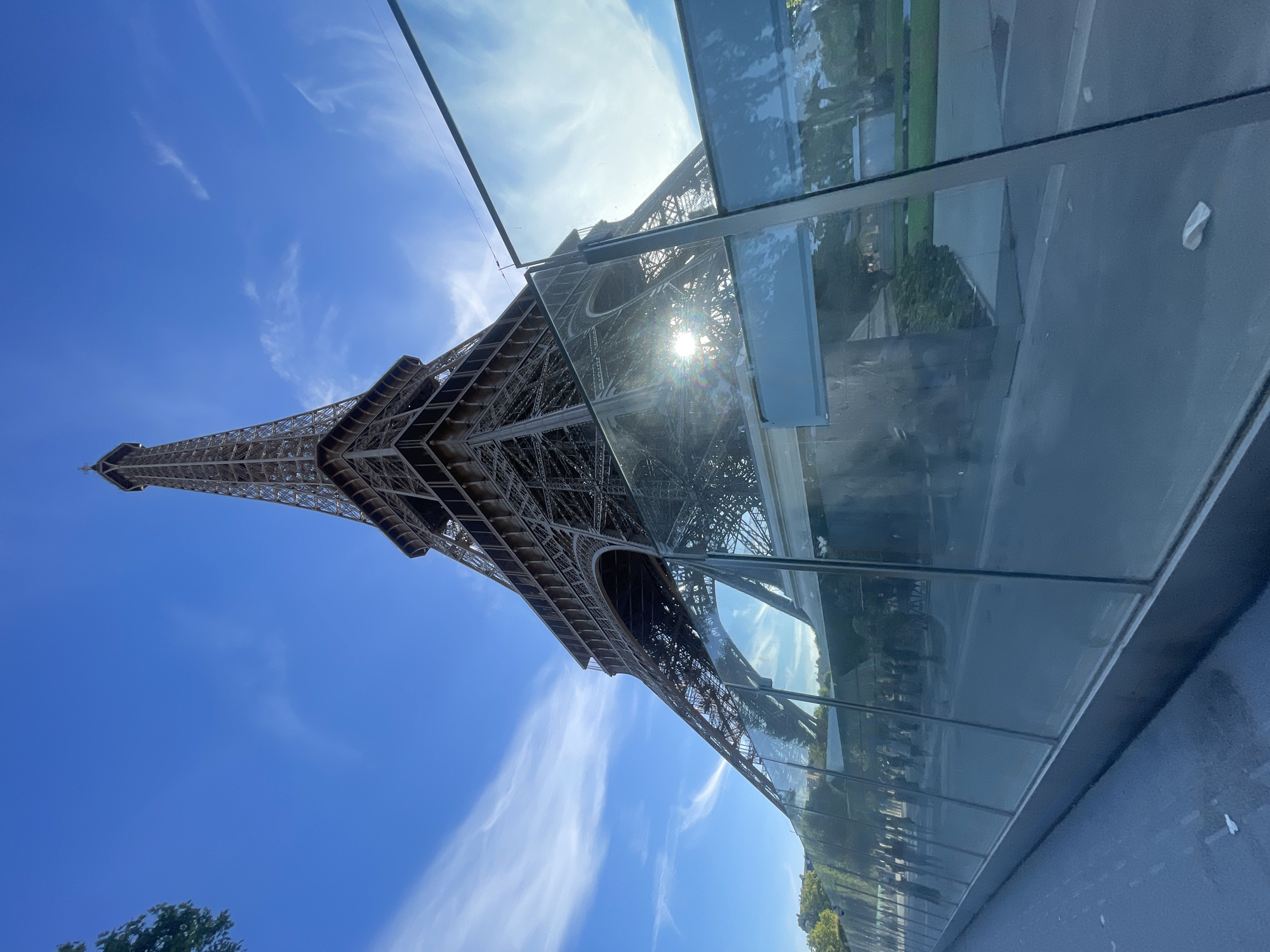 This media file captures a low-angle perspective of the Eiffel Tower in Paris, France. The iconic iron lattice structure dominates the frame, reaching towards a clear blue sky dotted with wispy white clouds. Sunlight streams through the tower, creating a lens flare effect. In the foreground, a modern glass barrier or structure is visible, reflecting the sky and the Eiffel Tower, creating an interesting interplay of real and reflected imagery. The glass barrier has a sleek, horizontal design with visible seams.
The scene appears to be set during daylight, likely late morning or early afternoon, given the bright sunlight and clear sky. The overall atmosphere is bright and sunny, suggesting pleasant weather. The presence of the Eiffel Tower and the general ambiance point to a tourist or public area in Paris. There are no people clearly visible in the shot, focusing the attention solely on the architecture and the sky. The ground visible at the bottom appears to be a paved area, possibly a walkway or road. The reflection in the glass shows glimpses of trees and what might be surrounding buildings or parkland. The image is oriented vertically, with the top of the Eiffel Tower pointing towards the bottom of the frame.