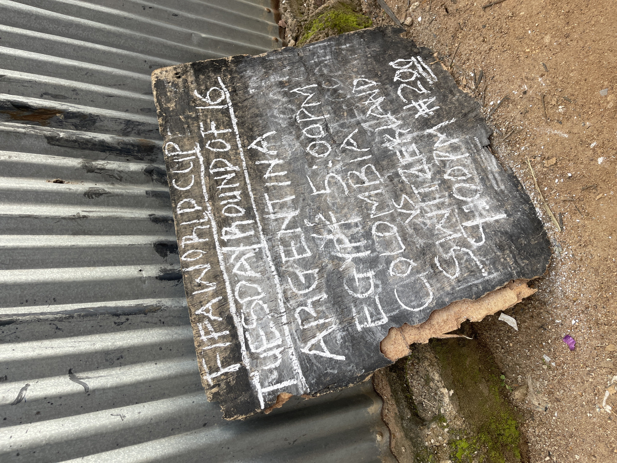 A rough-hewn, dark wooden plank, propped against a corrugated metal fence, displays handwritten white chalk lettering. The sign advertises "FIFA WORLD CUP" and "TUESDAY ROUND OF 16". Below this, it lists "ARGENTINA", followed by "EGYPT VS 5:00PM". The next line reads "COLOMBIA VS SWITZERLAND" and the final line indicates "4:00PM #200". The fence is weathered, with streaks of rust and dirt. The ground is a mixture of soil, small stones, and dried leaves, with a patch of green moss visible on the left. A small piece of purple plastic is on the ground in the foreground. The scene appears to be outdoors, likely in a somewhat informal setting. The location is identified as Rafin Par, Nigeria.