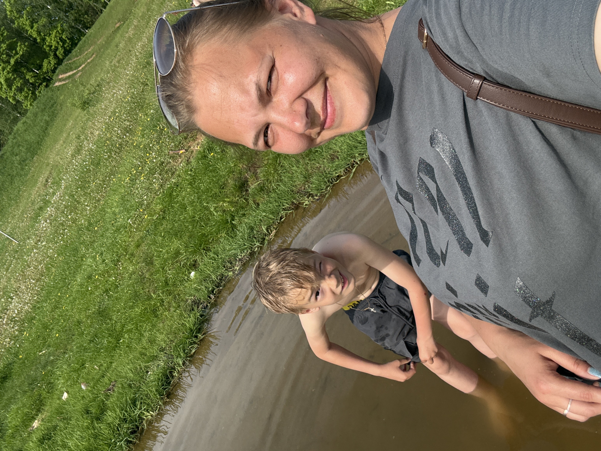 A woman and a boy are standing in a body of water with grassy shores in Novosinkovo, Russia. The woman is smiling and looking at the camera while taking a selfie. She is wearing sunglasses on her head, a gray T-shirt, and has a brown purse strap over her shoulder. The boy is wearing black shorts and a necklace with a cross. The water appears to be murky. The grassy shoreline is visible on the left of the frame.