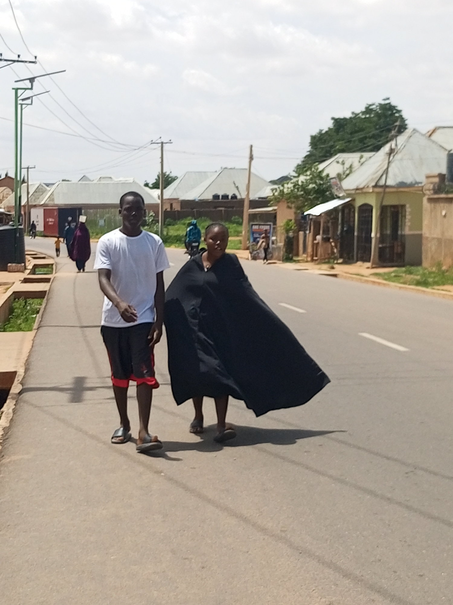 In Tempure, Nigeria, two individuals are walking on a paved road on a cloudy day. On the left, a man wearing a white t-shirt and black shorts with red trim walks towards the viewer. He is wearing dark flip-flops. To his right, a woman is walking in the same direction. She is wearing a flowing black garment that covers her from head to toe, obscuring her face and body. Her feet are visible, and she is wearing light-colored sandals. The road has a white dashed line marking its center. On the left side of the road, there is a raised sidewalk with some greenery. In the background, there are buildings with corrugated metal roofs and some trees. Power lines are visible overhead, supported by utility poles. Further in the background, a few other people and a motorcycle can be seen.