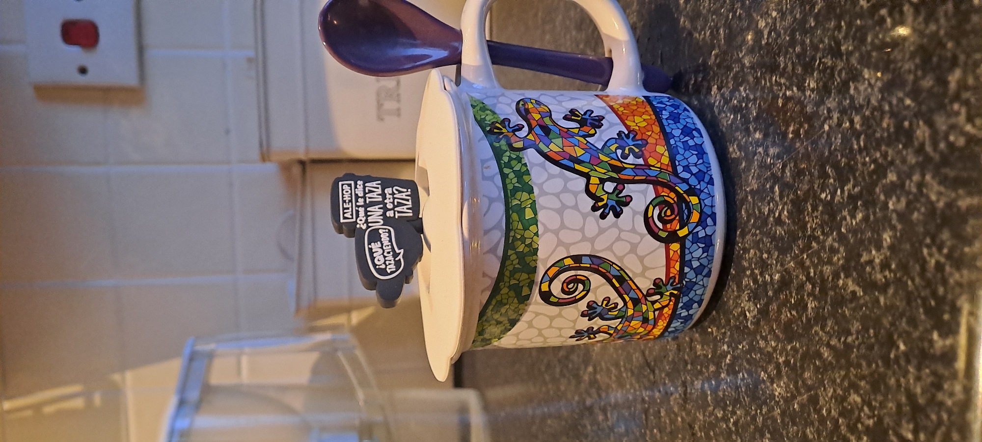 The image shows a close-up view of a mug with a lid and a spoon. The mug has a white lid with a black novelty charm attached, featuring text in Spanish and English. The mug itself is decorated with colorful mosaic-style lizards and abstract patterns. A purple spoon is inserted into the mug's rim. The mug sits on a dark, speckled countertop. In the background, out of focus, are kitchen cabinets and a light-colored wall with tiles. The overall scene appears to be indoors, likely in a kitchen setting. There are no people or animals present in the image. The location is not identifiable as Castlebar, Ireland; the image is of a domestic kitchen item.