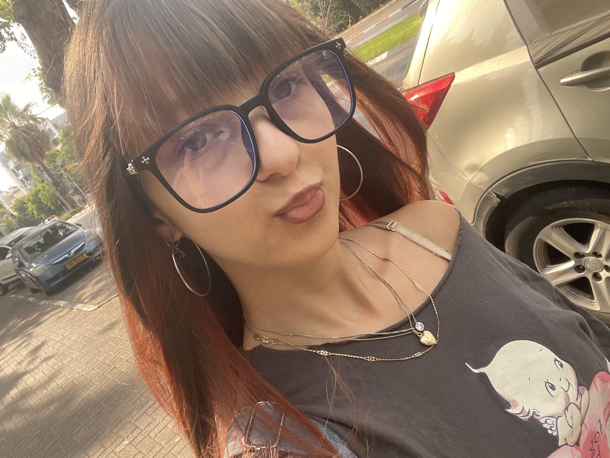 The image is a selfie taken from a high angle, looking down at the subject. The subject is a person with long, reddish-brown hair and bangs, wearing large, dark-rimmed glasses with silver accents. They are wearing a dark gray t-shirt with a graphic print of a baby on it, layered over a light-colored bra strap. Around their neck, they have multiple layered necklaces, including a gold chain with a small heart pendant and a diamond accent. They are also wearing large hoop earrings.
The background suggests an urban setting, likely in Raanana, Israel. A paved sidewalk with a patterned texture is visible in the upper left, with shadows cast on it, indicating sunlight. A dark-colored car is parked on the street. Further in the background, palm trees and buildings are visible, consistent with a city in Israel. To the right of the subject, the rear of a light-colored car, possibly a champagne or gold SUV, is visible. The car's taillight is illuminated red, and its wheel and part of the fender are in view. The overall lighting suggests it is daytime, with bright sunlight.