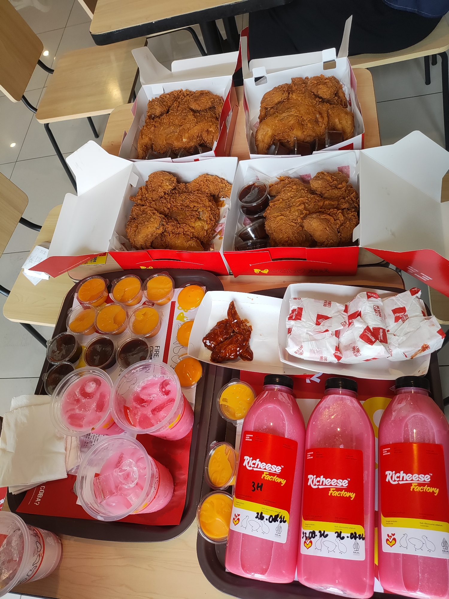 The image captures a tabletop arrangement of fast food from Richeese Factory. In the background, two open boxes of fried chicken are visible, each containing several pieces of golden-brown fried chicken. In the mid-ground, a dark tray holds an assortment of food items. This includes several small plastic cups filled with an orange-colored sauce, and others with a dark brown sauce, likely dips for the chicken. There are also three clear plastic cups filled with a pink, ice-filled beverage. Further to the right on the tray, a small white box contains a few pieces of glazed chicken wings, and another white box is filled with individually wrapped items, also from Richeese Factory. In the foreground, three large pink plastic bottles with black caps, also bearing Richeese Factory branding, are lined up. The setting appears to be an indoor seating area, possibly a restaurant or a dining hall, with wooden tables and chairs visible in the background. Based on the context and branding, this scene is likely in Majalengka Kulon, Indonesia, a location known for its fast-food establishments.