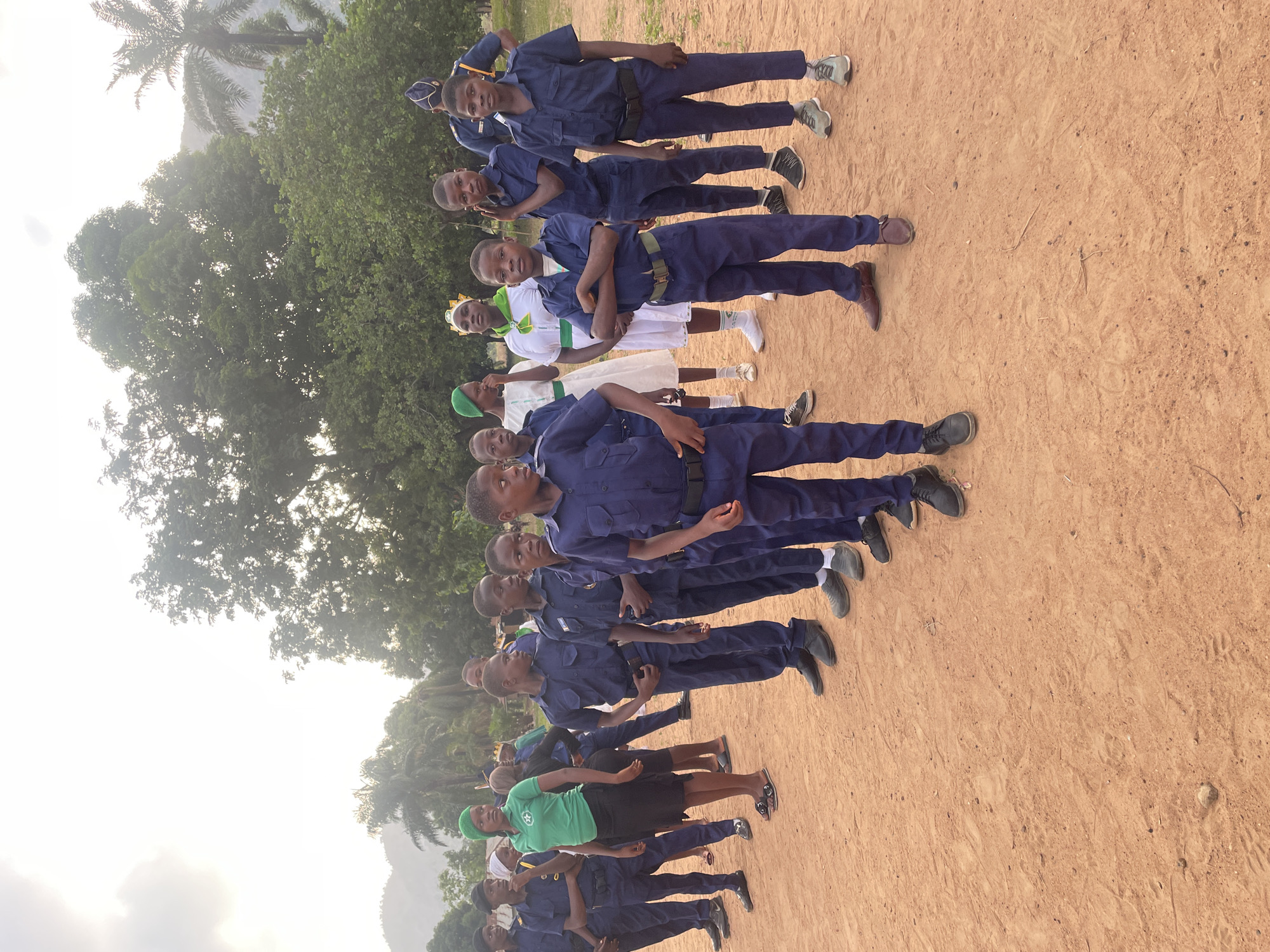 A group of approximately 15 people, mostly young males, are standing in rows on a dirt ground in Andaha, Nigeria. The majority of the group is wearing matching dark blue uniforms with belts and dark shoes. Several individuals in the middle rows wear white uniforms with green sashes and green head coverings. They all appear to be standing at attention. The environment includes green trees, a palm tree, and possibly a mountain in the background under a slightly overcast sky.