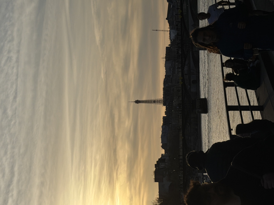 The image displays an urban riverfront scene in Paris, France, captured at sunset or dusk. The Eiffel Tower stands centrally in the far background, silhouetted against a cloudy sky that shifts from a golden-orange hue on the left horizon to a paler grey-blue overhead. The Seine River occupies the mid-ground, traversed by a multi-arched bridge with visible piers. A dense cityscape forms the backdrop behind the bridge, featuring buildings and a tall construction crane visible on the far right. In the immediate foreground, a riverside promenade is populated by multiple individuals. A person with long dark hair, wearing a dark top, is partially illuminated while appearing to walk or jog forward on the right. To their left, another individual is seen from behind, silhouetted, with headphones visible. Additional silhouetted figures are present, some standing or seated along a dark metal railing that separates the promenade from the river.
