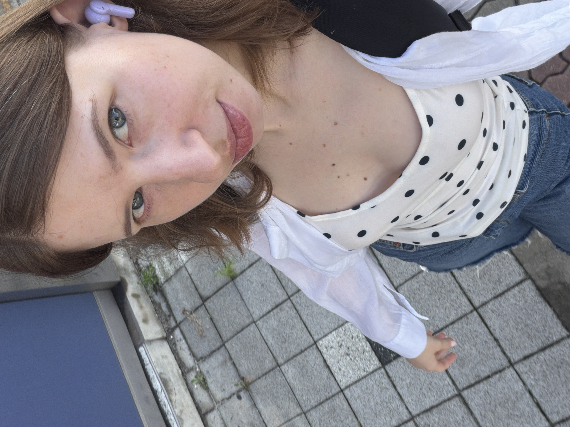 A young woman is taking a high-angle selfie outdoors in Dongducheon-si, South Korea. She has brown hair and blue eyes and is wearing a white collared shirt, a white polka dot tank top, and blue jeans. She has a purple wireless earbud in her right ear. The background consists of a tiled sidewalk and a blue wall.