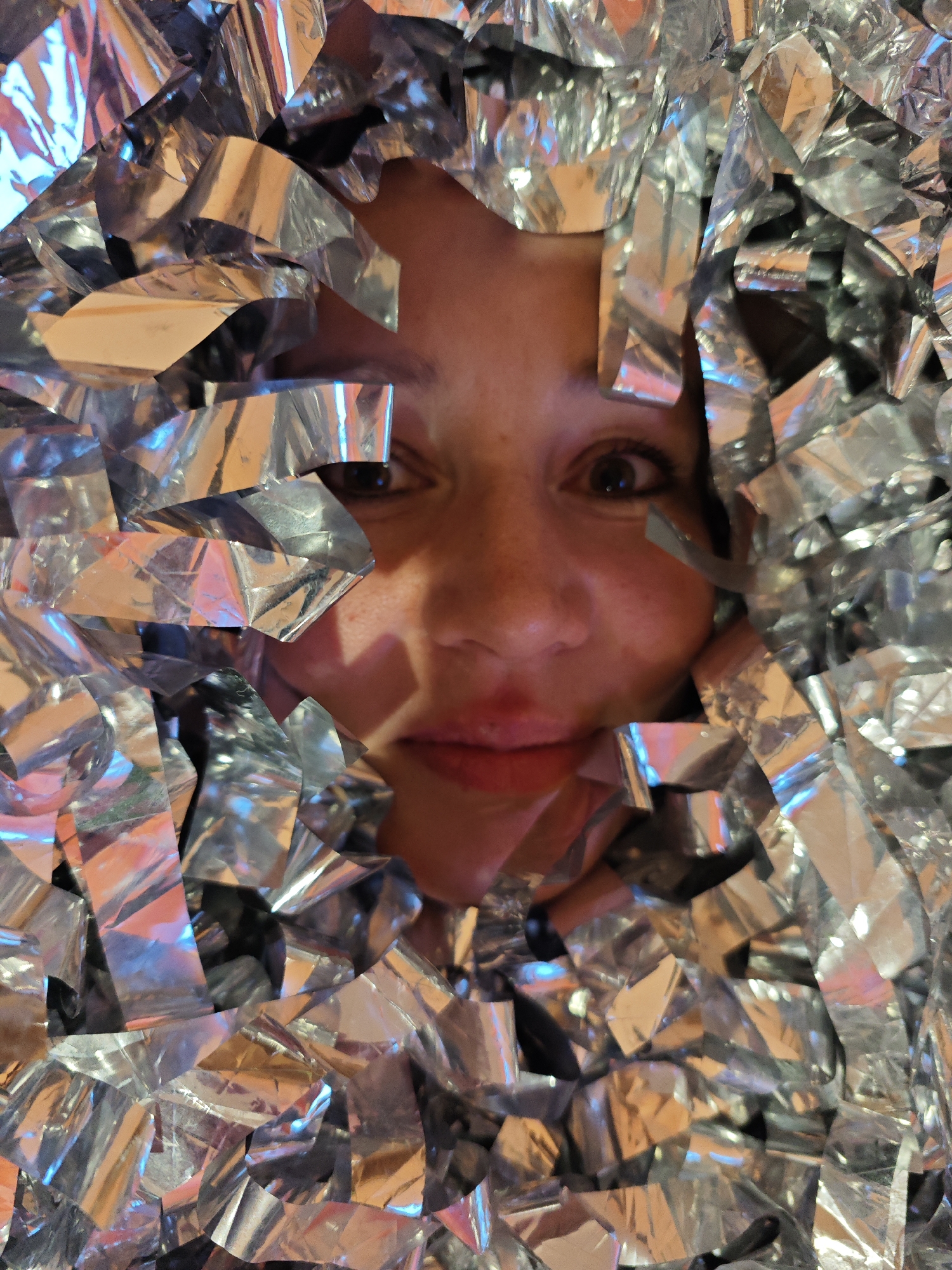A person's face is visible through a mass of shiny, silver confetti. The face is fair-skinned with freckles and has a slight smile. The person's eyes are open and looking directly at the viewer. The confetti is irregularly shaped and highly reflective, catching different colors of light. The background is obscured by the mass of confetti. There is no evidence to support the claim that this image was taken in Markovo, Russia.