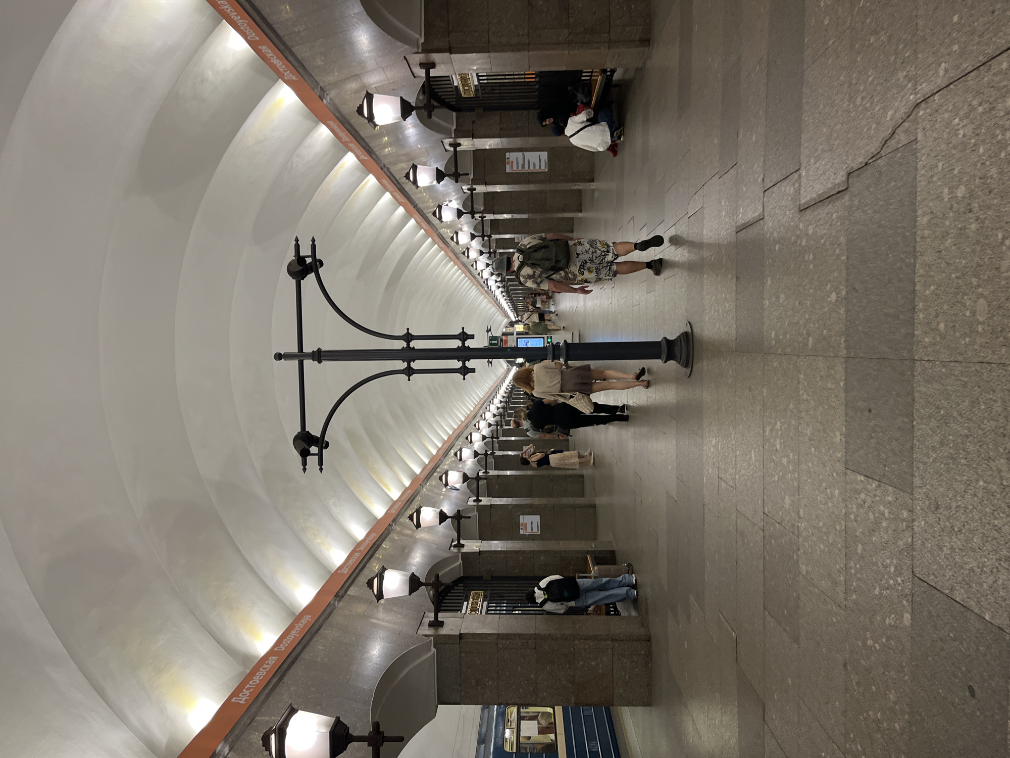 The image shows the interior of the Dostoyevskaya metro station in Saint Petersburg, Russia. The station features a vaulted ceiling with recessed lighting and decorative lamps lining the platform. The platform is paved with large granite tiles. Several people are visible on the platform, some walking and others sitting on benches. A blue metro train is stopped at the platform on the left side of the image. The walls are covered with granite panels, and orange signage with the station name in Russian and English is present along the upper part of the walls. The overall scene depicts a busy public transportation environment.