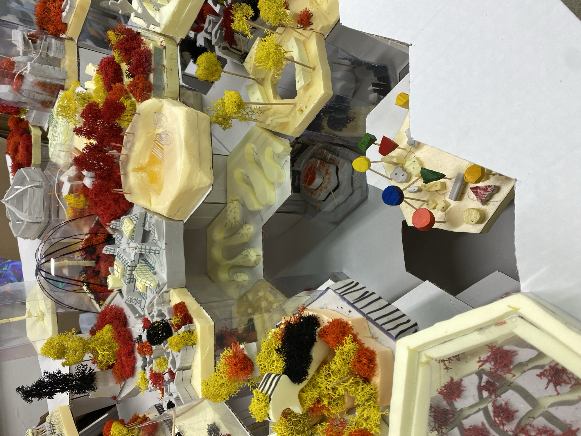 The image shows a detailed architectural model, constructed with hexagonal foam cells. The cells contain various elements including miniature trees (yellow and orange), abstract white shapes, and small colorful wooden pieces. Some cells are covered with clear plastic, providing a glimpse into the contents. The model seems to represent an abstract urban landscape or architectural concept. There is no evidence to indicate the location as Moscow, Russia.