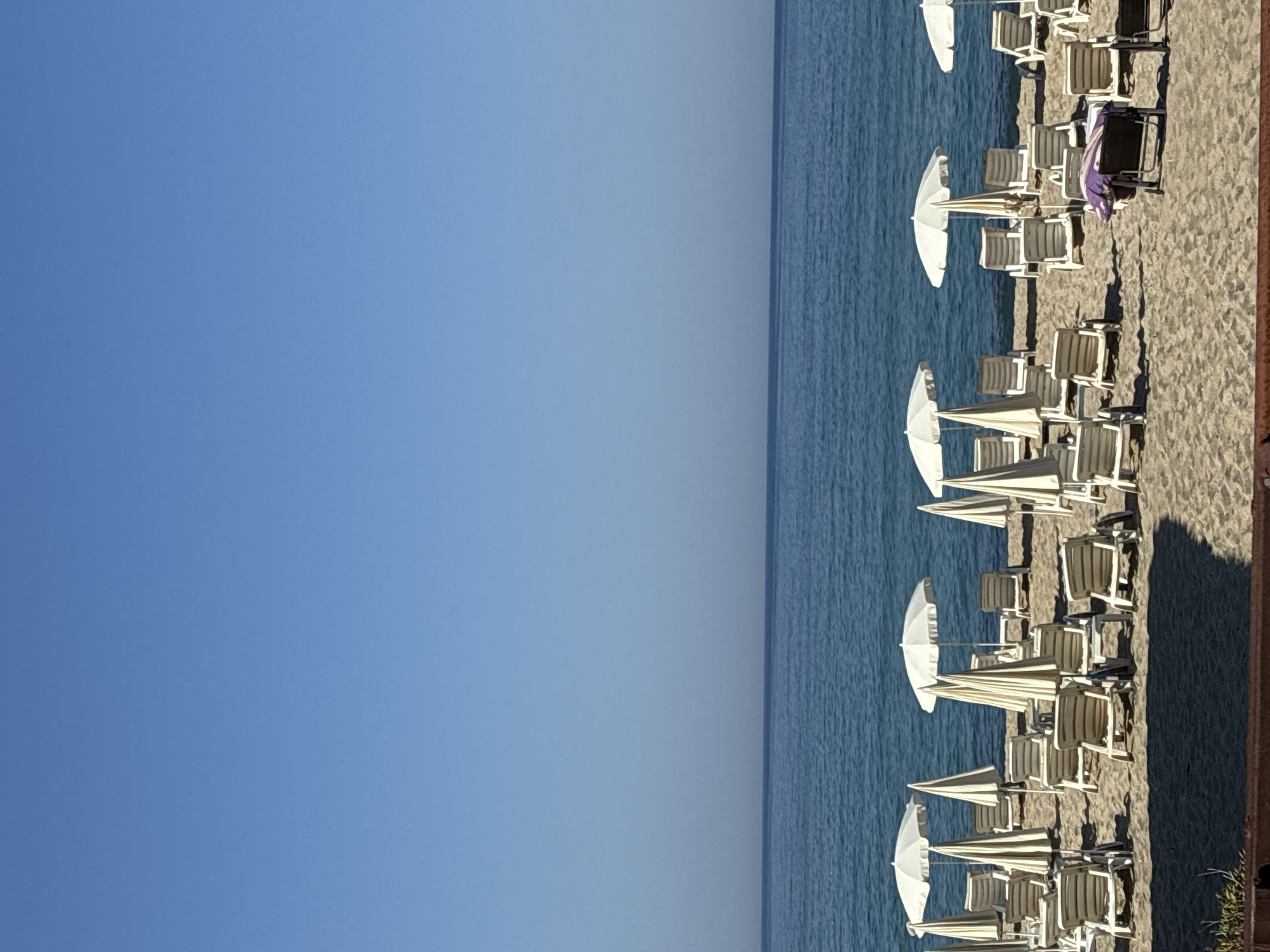 The image depicts a beach scene in Alanya, Turkey, with rows of empty lounge chairs and white umbrellas set up on the sand. The sea is visible in the background, stretching out to the horizon under a clear blue sky. The beach appears to be unoccupied, with no people present. The overall impression is one of tranquility and readiness for visitors, typical of a well-maintained tourist beach.