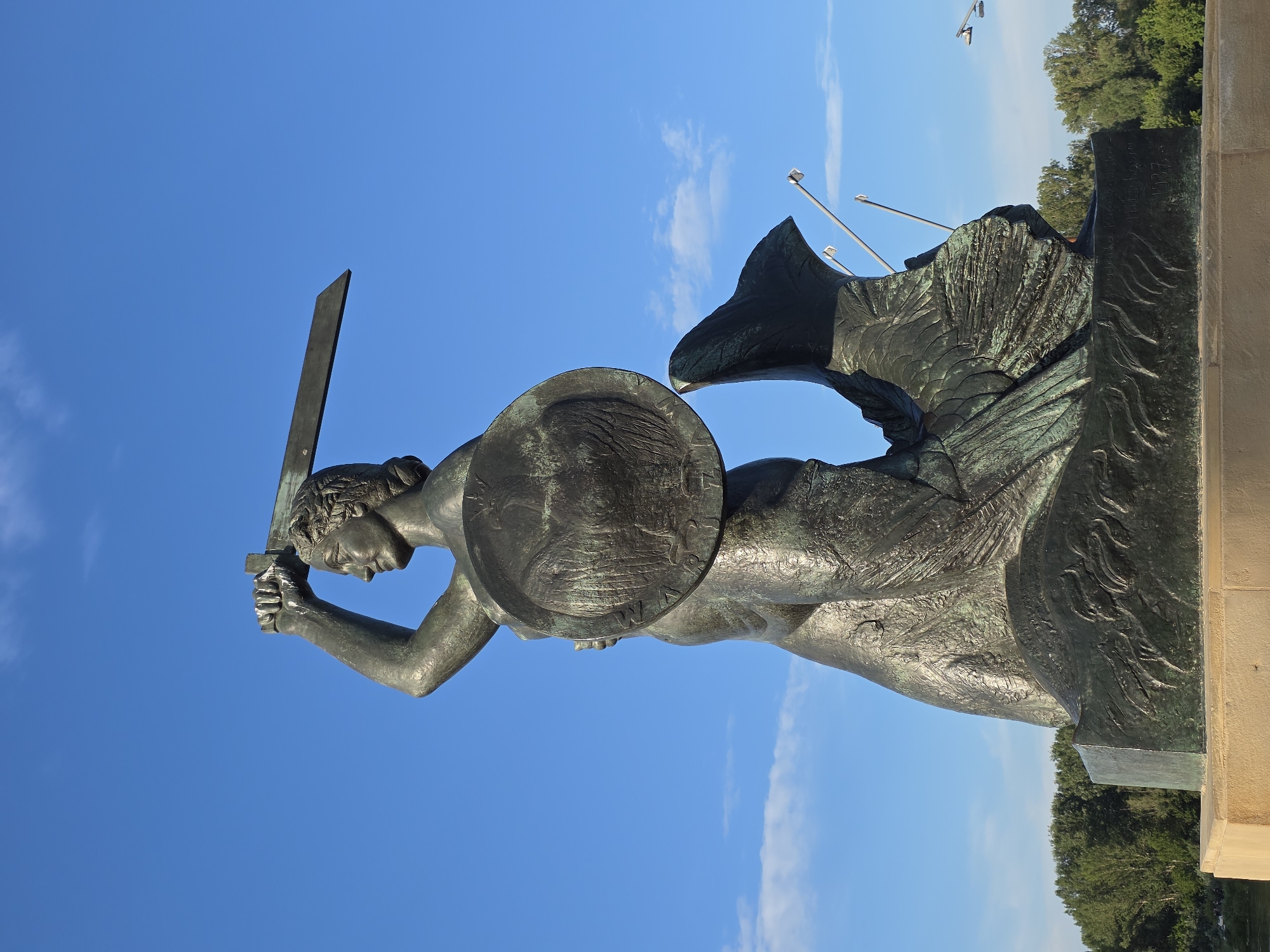 The image depicts the Warsaw Mermaid statue, a bronze monument located on the Vistula River embankment in Warsaw, Poland. The statue, facing to the left, is shown from the chest up against a clear blue sky with sparse white clouds. It portrays a mermaid holding a sword aloft in her right hand and a shield in her left. The mermaid has a muscular build and her lower body is depicted as a fish's tail. The bronze has a weathered patina, with hints of green. The statue is mounted on a stone pedestal. In the background, a line of green trees is visible, suggesting an outdoor setting, possibly a park or riverside area. Several streetlights and flagpoles are also present in the distant background. The lighting suggests it is daytime.