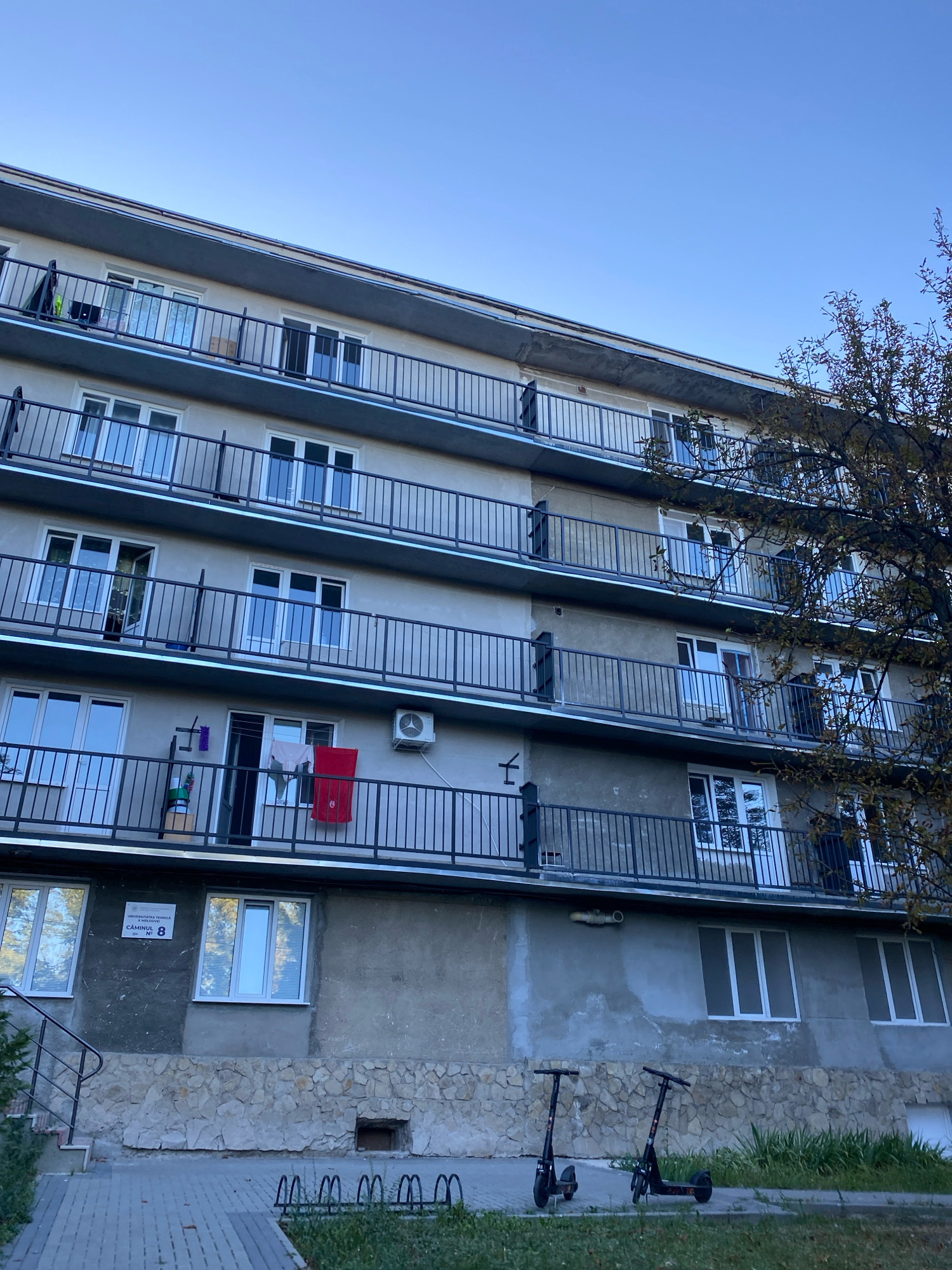 The image shows a side view of a multi-story apartment building with concrete walls and balconies on each floor. The building has a gray facade with numerous windows and black railings on the balconies. Laundry is visible hanging from a balcony on the fourth floor. To the right of the building, a tree with autumn foliage is partially visible. In the foreground, there is a paved walkway and a grassy area with two electric scooters parked. A sign on the building, visible in the lower left, reads "UNIVERSITATEA TEHNICĂ CHIȘINĂU N° 8". This indicates the location is Technical University Dormitory No. 8 in Chișinău, Moldova. The sky is clear and blue, suggesting it is daytime. There are no people visible in the image.