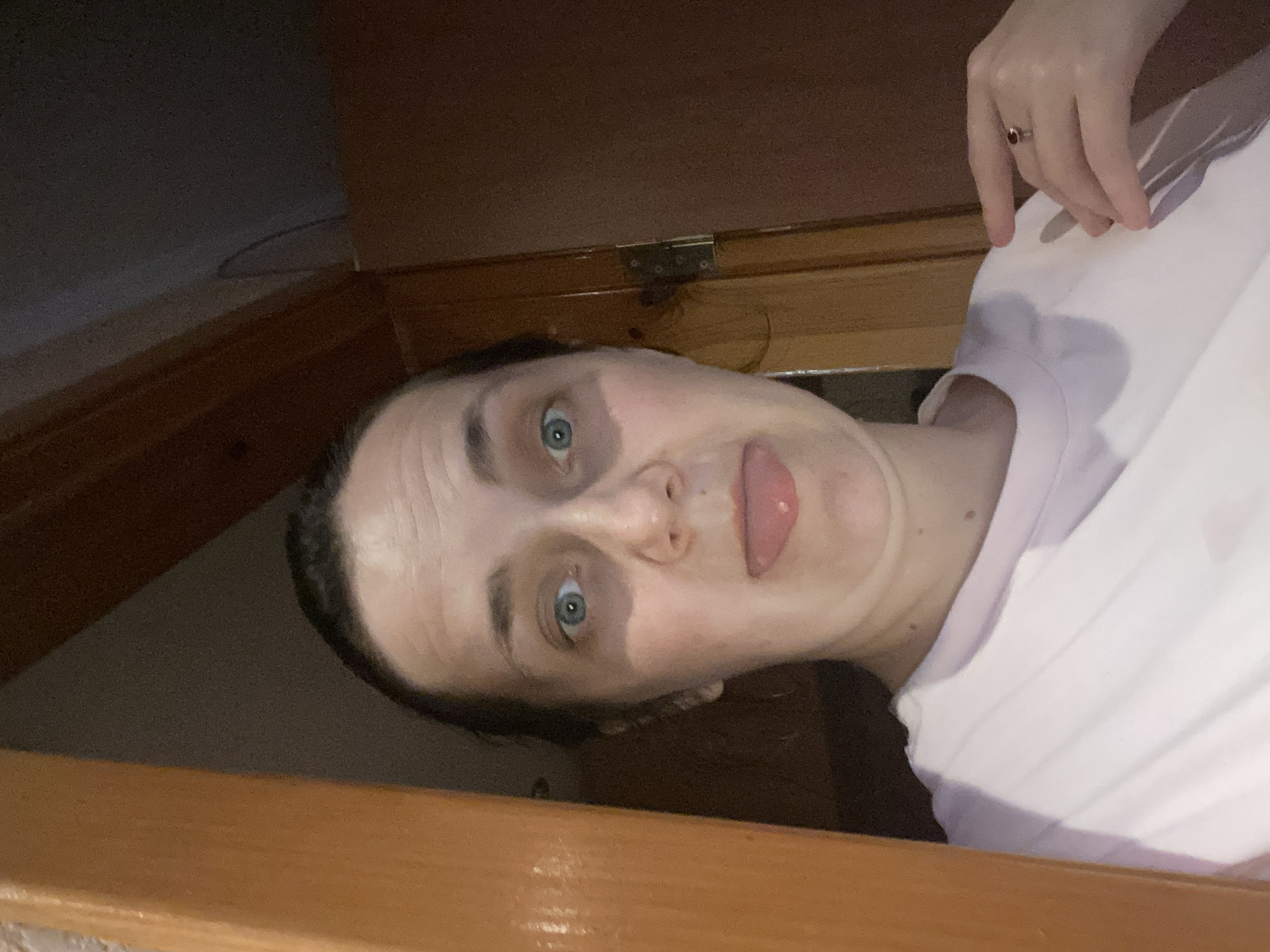 A medium shot captures a woman from the chest up, positioned in what appears to be a doorway. She is wearing a pale pink t-shirt and her left hand, adorned with a ring on the index finger, rests on her shoulder. Her head is turned slightly towards the camera, with her tongue playfully sticking out. The background consists of a wooden doorframe and a dark wall. The lighting is dim, suggesting an indoor setting at night or in a low-light environment. The woman's expression is one of lightheartedness and playfulness.