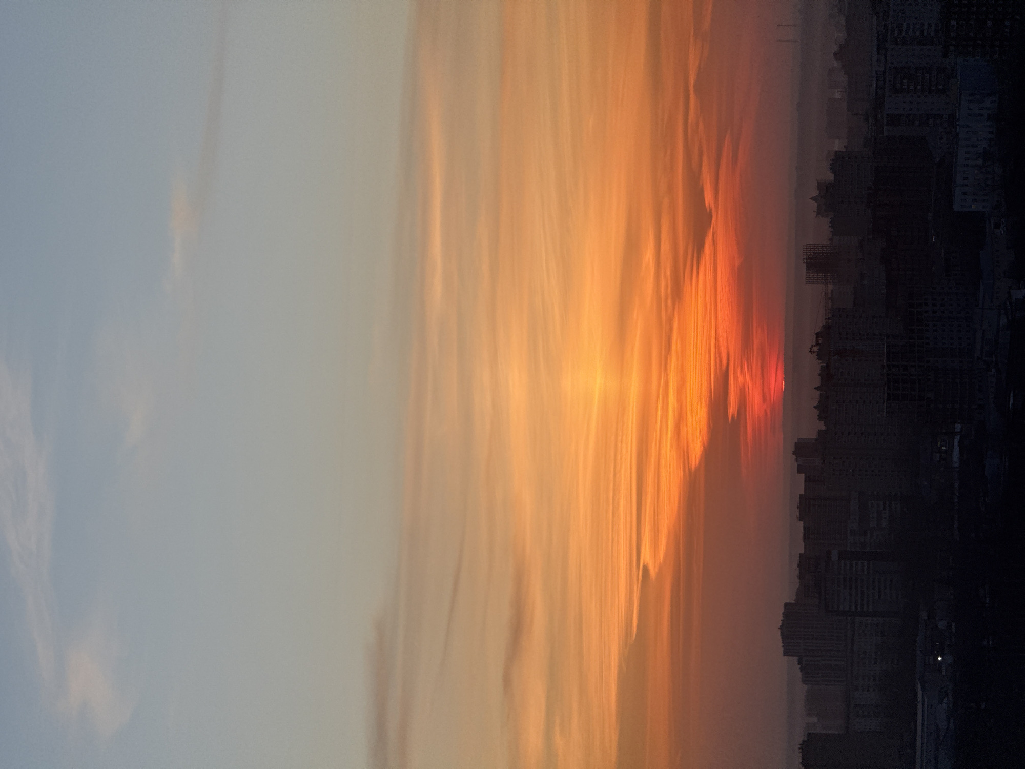 The image depicts a sunset over the city of Perm, Russia. To the right is the silhouetted skyline of the city, including several high-rise buildings. The sunset is a vibrant mix of colors, with orange and red hues dominating the sky above the buildings. The left side of the image shows the sky fading to a light blue color. The image is rotated 90 degrees counter-clockwise. There are no people visible in the image.