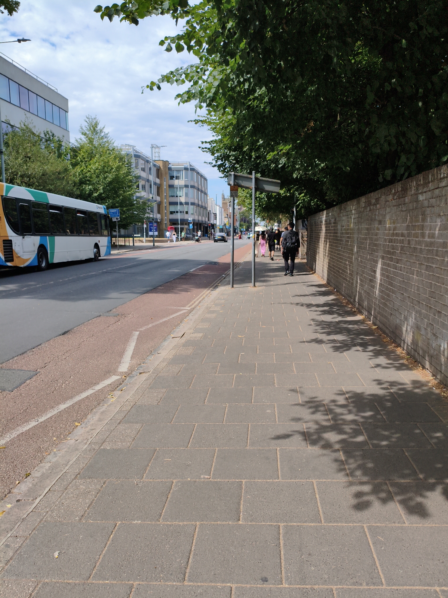 The image depicts a street scene in Cambridge, United Kingdom, likely on a summer day with a bright, partly cloudy sky. A white bus with colorful stripes is parked on the left side of the road. The bus appears to be a public transport vehicle, possibly a city bus. Adjacent to the road is a sidewalk paved with large, rectangular grey tiles.
Several people are walking along the sidewalk. In the foreground, a person with a backpack is walking away from the camera. Further ahead, a group of people, including at least one person in a pink garment, are also walking. The sidewalk is bordered on the right by a brick wall. The wall is constructed from light brown bricks and shows some signs of weathering. Above the wall, the lush green foliage of trees overhangs the scene, casting dappled shadows onto the sidewalk.
Behind the sidewalk and bus, there are modern buildings with glass facades and some older brick structures, typical of an urban environment. Traffic signals are visible in the distance, indicating this is a functioning street with vehicular traffic. The road itself is asphalt, with a red-colored lane marker for a bus lane or cycle path. A street lamp is visible on the left side of the frame. The overall impression is of an everyday urban scene in Cambridge.