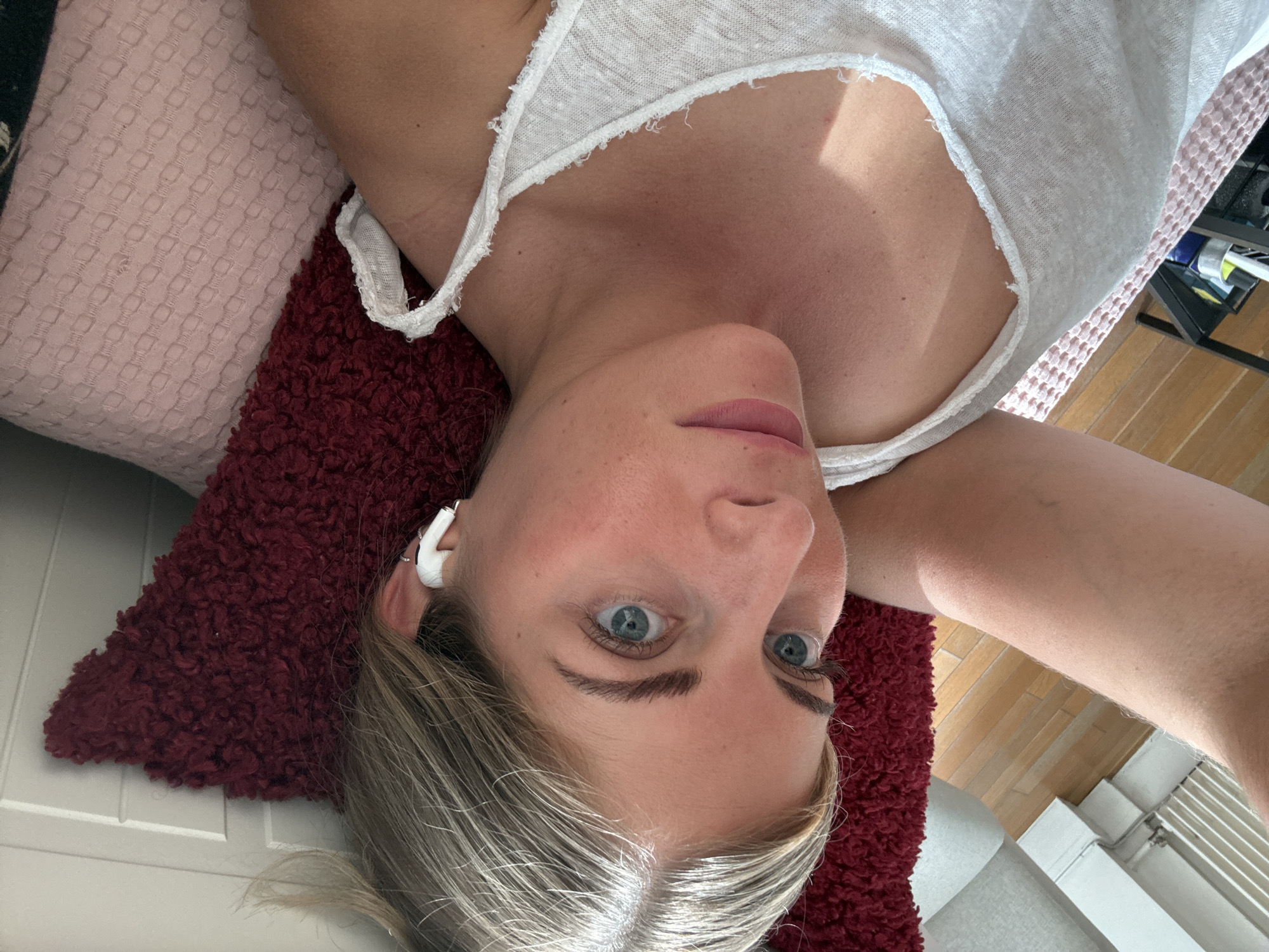 A person lies on a bed in a room with hardwood floors and white walls. The person, presumably female, has light skin, light hair, and blue eyes. She is wearing a white top and a white earbud is visible in her left ear. Her head rests on a red, textured pillow, which is in turn on a pink patterned blanket. A radiator is visible in the lower right corner of the frame. The location cannot be confirmed.