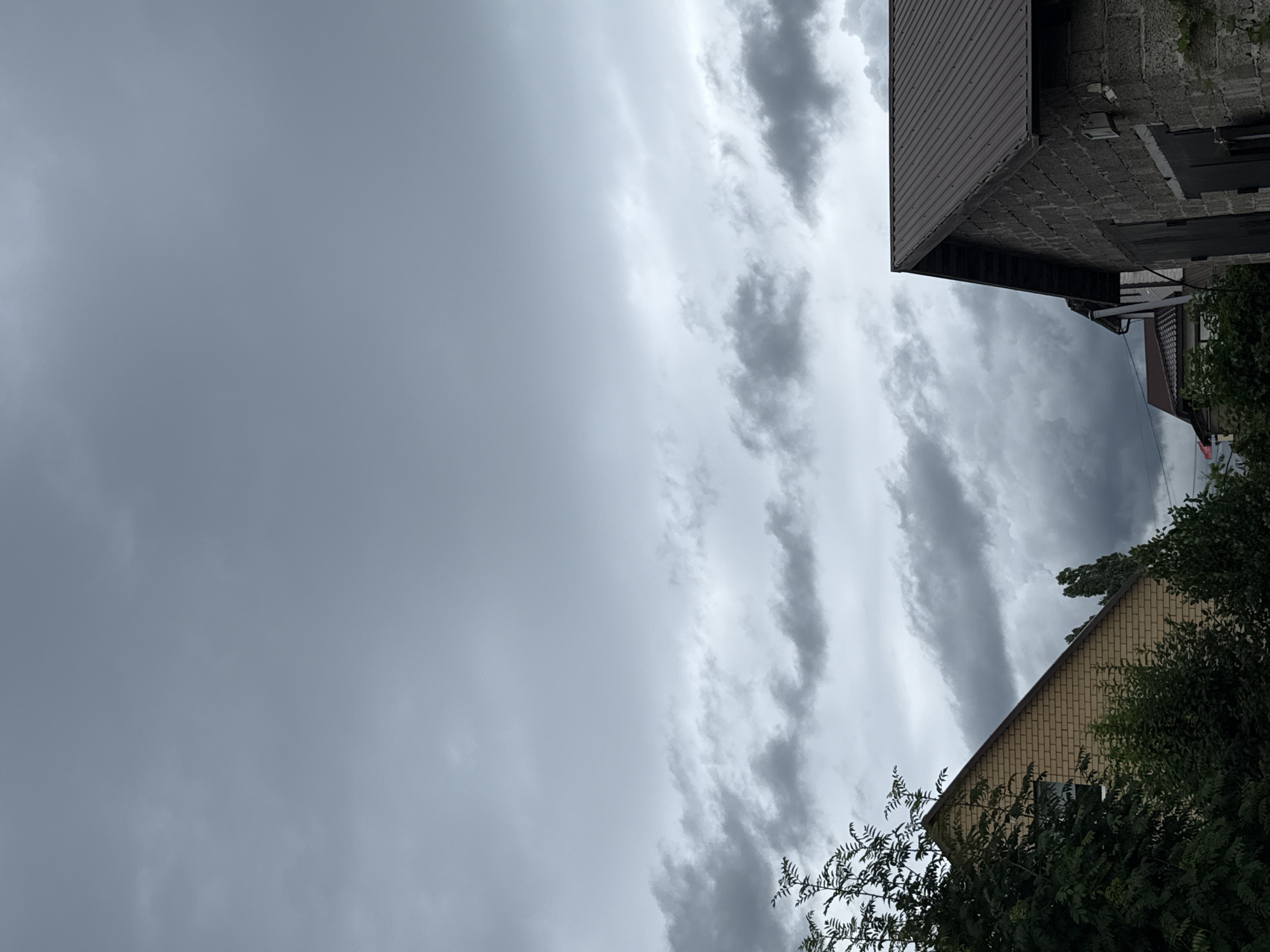 The image depicts a cloudy sky over a residential area. In the foreground, on the right, is the corner of a dark-roofed building constructed from gray cinder blocks. To the left of this building, a portion of another building with a lighter-colored brick facade and a dark roof is visible. Dense green foliage obscures the lower parts of both buildings. The sky is filled with overcast, gray clouds, with lighter patches suggesting diffused sunlight. The scene appears to be a residential street or backyard. No people or specific actions are identifiable in the image.