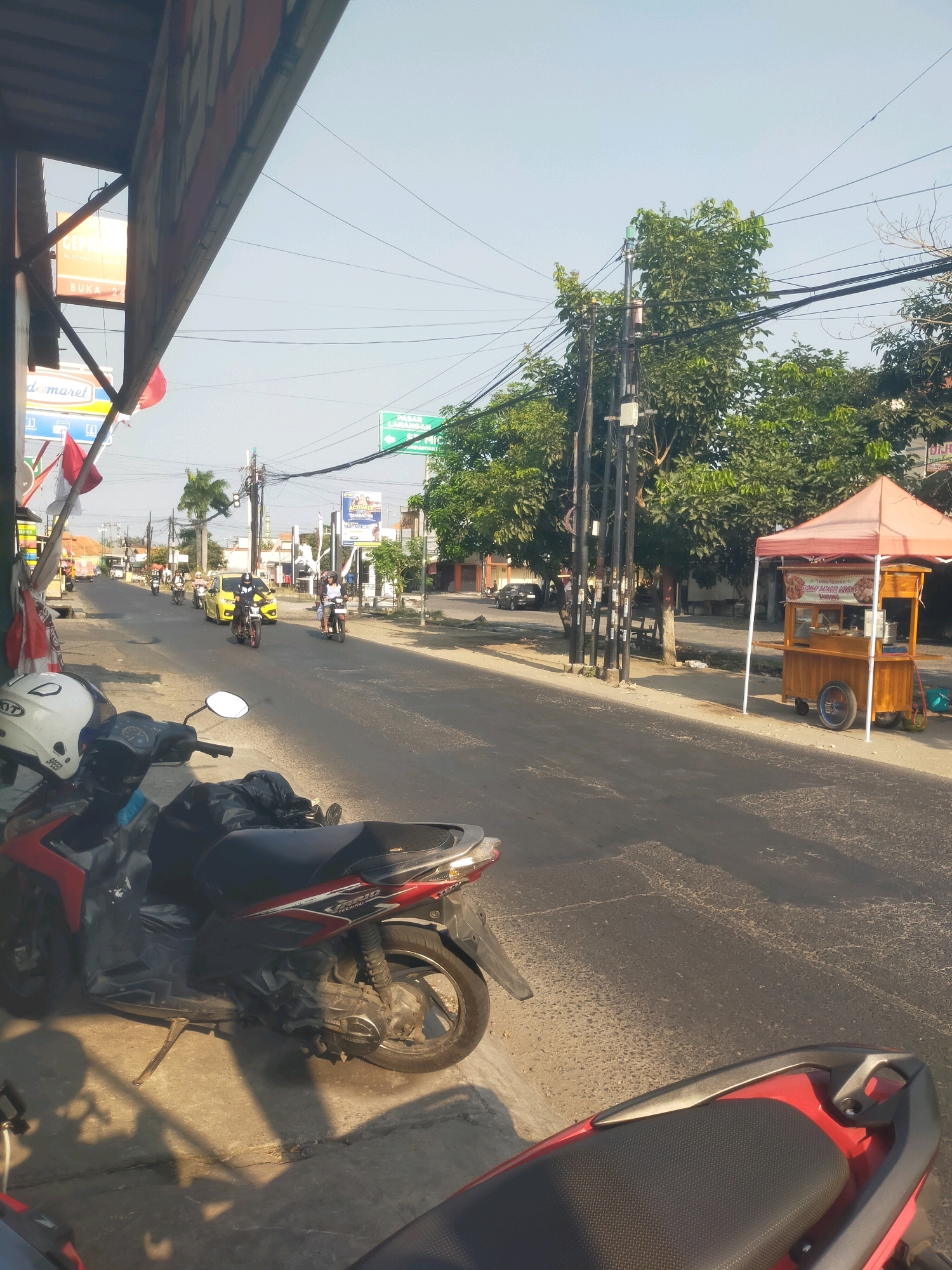 In the city of Sidodadi, Indonesia, a street scene unfolds with a clear sky overhead. The foreground is dominated by the right side of a parked red and black motorcycle, its handlebars and part of the seat visible. A white helmet rests on the left handlebar. Adjacent to the motorcycle, on the concrete sidewalk, shadows of foliage are cast.
The street itself is paved, with a bright yellow car and several motorcycles carrying riders traveling away from the viewer. Buildings line the right side of the street, and a food cart with an orange canopy and wooden structure is set up by the roadside. The cart displays signage and appears to be selling food. Trees with dense green foliage are visible behind the cart and further down the street, interspersed with utility poles and electrical wires.
A blue and white sign with "Indomaret" is visible on the left side of the street, indicating a convenience store. Other signs, including a green directional sign, are present further down the road. The overall impression is of a typical, sunny day on a street in a populated area, likely a commercial or residential district.