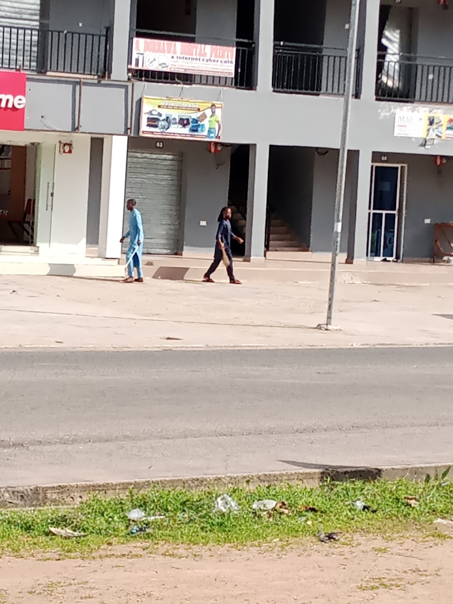 The image depicts a street scene in Jalingo, Nigeria, likely in front of a commercial building. Two individuals are walking on the sidewalk. The person on the left is wearing a light blue garment, and the person on the right is wearing a dark blue garment. The building behind them has multiple levels with balconies and shopfronts. One shop displays signage with text that includes "A BUKANA DIGITAL PRINTING" and "A Internet cyber cafe." Another sign further down the building reads "AFAS." The street in front of the building is paved, and a section of a grassy embankment with scattered litter is visible in the foreground. A lamppost stands on the sidewalk. The lighting suggests it is daytime.