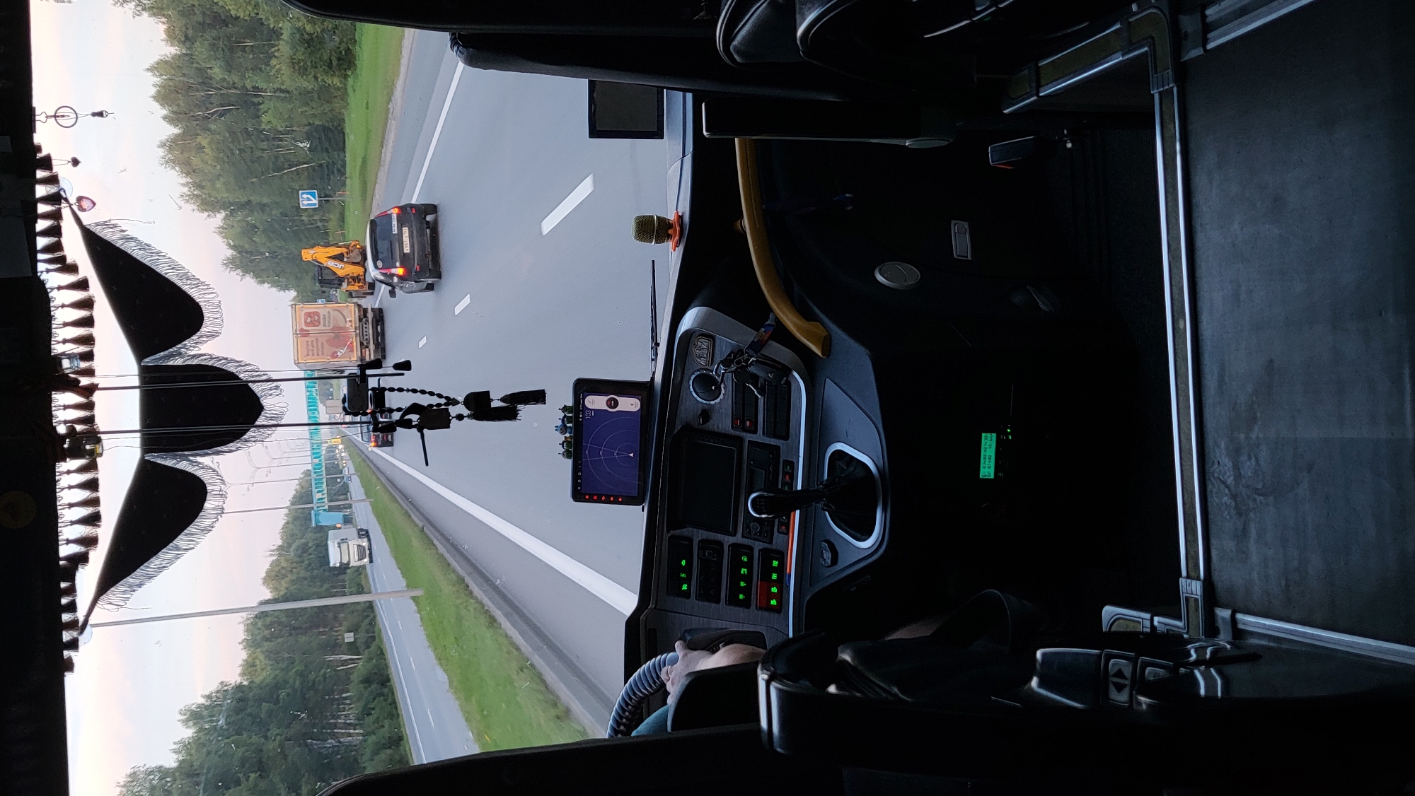 The image is taken from the interior of a bus or truck, looking forward through the windshield. The view shows a highway with multiple lanes of traffic, flanked by trees. Several vehicles are visible on the road, including a white truck, a yellow excavator, a light-colored car, and a dark-colored car.
The interior of the vehicle features a dashboard with various controls, a gear shifter, and a GPS device mounted on the windshield. Decorative tassels and trinkets are hanging from the top of the windshield.
The image appears to have been taken on a highway in or near Gruzinskii, Russia, based on the road signs and the style of vehicles. The lighting suggests it is either early morning or late afternoon. There is no clear indication of people in the immediate foreground, other than a hand on the steering wheel. The environment is that of a transportation route.