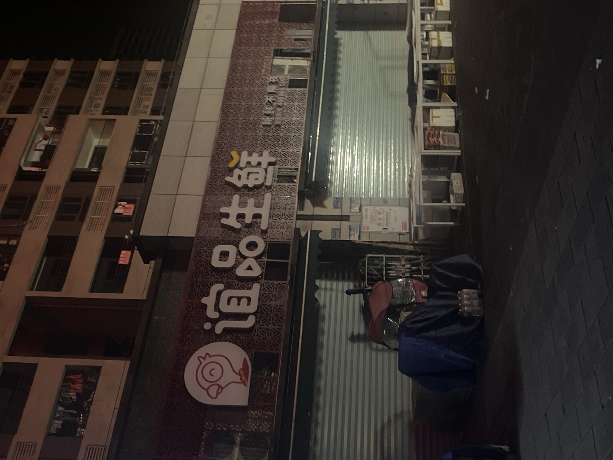This night-time image captures a quiet urban street scene in Chongqing High-tech Zone, China. The primary focus is a commercial shopfront, likely closed for the evening, indicated by a metal roller shutter door pulled down. The shop's facade features a prominent sign with Chinese characters "道品生鲜" (Dào Pǐn Shēng Xiān), which translates to "Dao Pin Fresh/Produce," suggesting it is a grocery or fresh food store. Below the text is a distinctive white logo depicting a stylized chicken or bird head.
To the left of the shop, a multi-story building with numerous windows and balconies extends upwards, appearing to be a residential or mixed-use structure. Adjacent to the shop's shuttered entrance, a metal shelving unit is visible, containing various stacked boxes and packaged goods, possibly supplies or deliveries. In the foreground, on the dark, paved sidewalk, a motor scooter or e-bike is parked, partially covered by a dark blue tarp, with its red seat and handlebars visible.
The scene is illuminated by artificial light sources, casting reflections on the roller shutter and highlighting elements of the buildings and sidewalk. The dark sky and the absence of people confirm the late hour. The architecture and vehicle are typical of an urban environment in China.