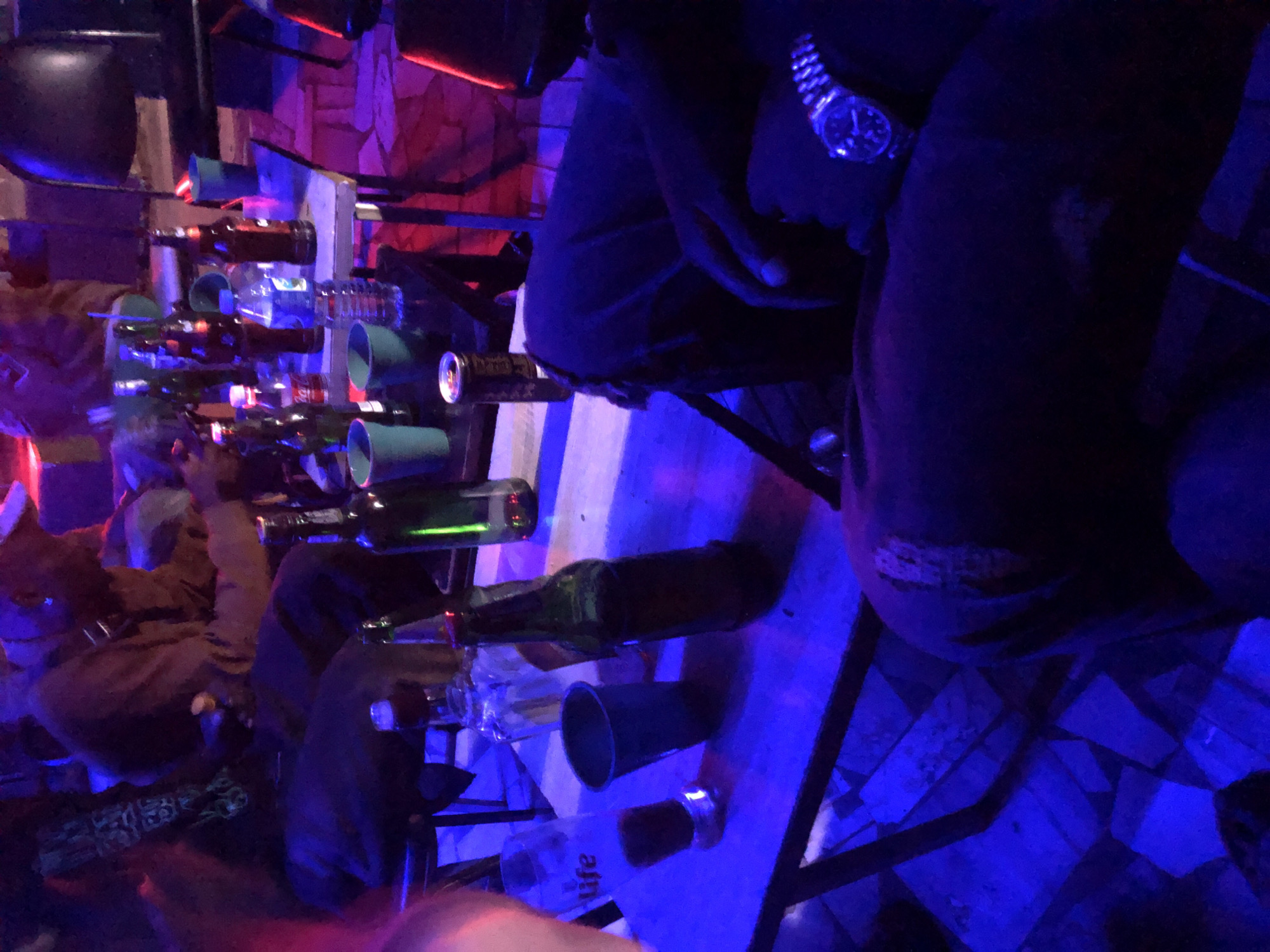 The image depicts an indoor social setting, exact city and country unknown. The scene is illuminated primarily by strong blue and purple artificial light, with some red light visible in the background, creating a low-light, atmospheric environment.
At least four individuals are visible. On the far left, a person's upper arm and shoulder are partially visible. In the mid-ground on the left, a person is seated, wearing dark trousers and a dark top with a visible pattern on the sleeve. A second person is seated in the mid-ground center-right, wearing a brown or tan top and a light-colored cap, looking downwards. In the foreground on the right, the lower body of a seated person is prominently featured, wearing dark trousers with a distressed area on the right knee. This person's right wrist displays a metallic wristwatch, and their left hand rests on their left thigh. Other indistinct figures are visible in the background.
Multiple tables are present. A light-colored rectangular table in the mid-ground holds several items, including numerous dark glass bottles (some green, some brown, consistent with beer bottles), a clear plastic water bottle, a small glass bottle with a red label (Coca-Cola), and a red-labeled can. Two green or teal-colored plastic cups are also on this table. A darker, smaller table in the foreground holds a dark-labeled can and another green or teal plastic cup. A clear glass labeled "Life" containing an amber liquid is also on this foreground table, alongside another dark glass bottle.
The floor is composed of light-colored, irregularly shaped tiles. Dark furniture components, likely chairs and table legs, are visible throughout the frame. A large, dark, rounded object, possibly a speaker or decorative fixture, is in the upper right background. Individuals are seated around tables, implying a social gathering, with various beverages present for consumption. No explicit interactions between individuals are discernible.