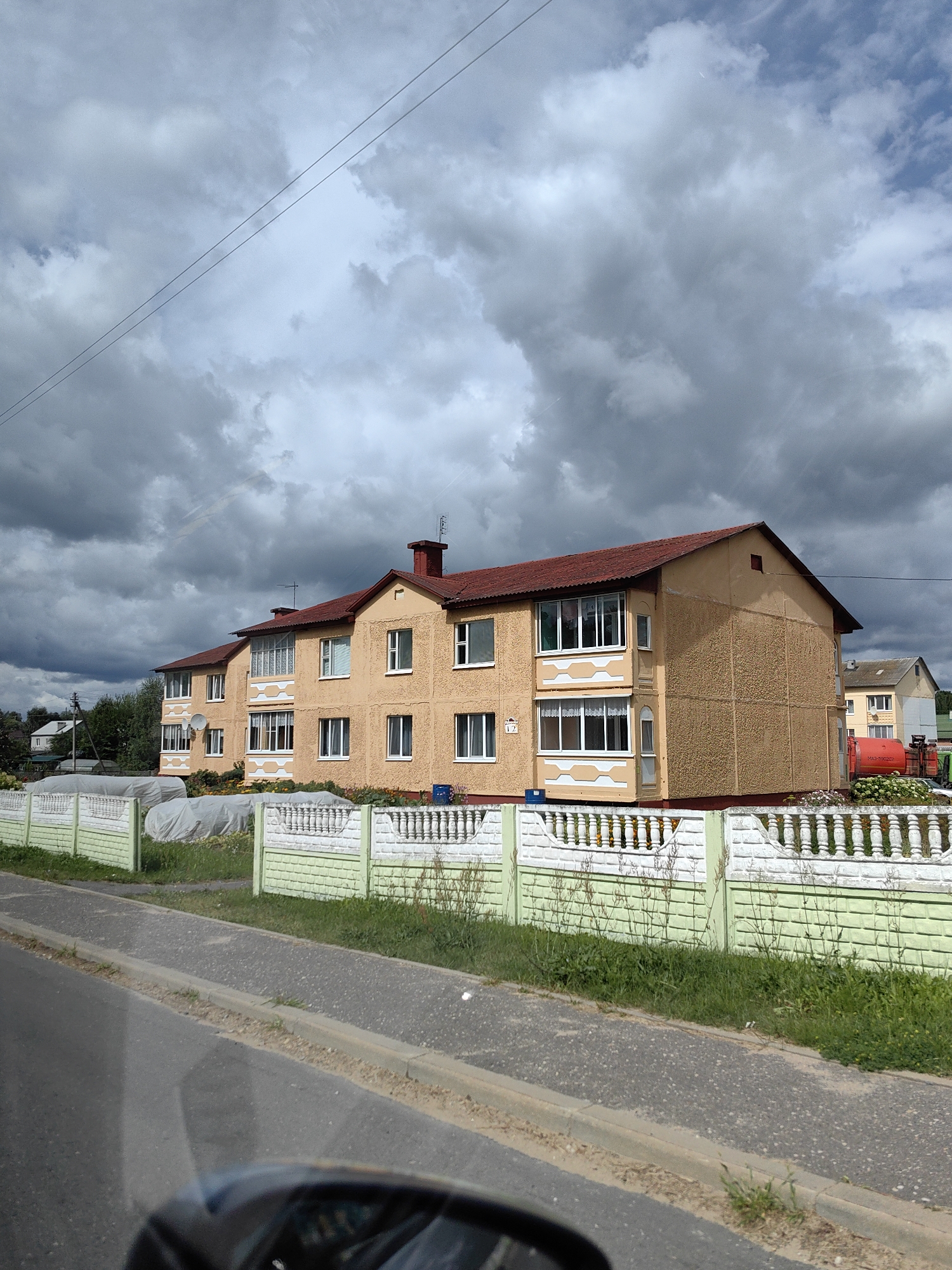 The image depicts a yellow, multi-story apartment building with a red tiled roof, located in Myadzyel, Belarus. The building is textured and has several windows and balconies. In front of the building, there is a decorative fence, a grassy area, and a road. The sky is cloudy and dark, suggesting an overcast or stormy day. A red tanker truck is parked in the yard behind the fence. The scene appears to be a residential area.