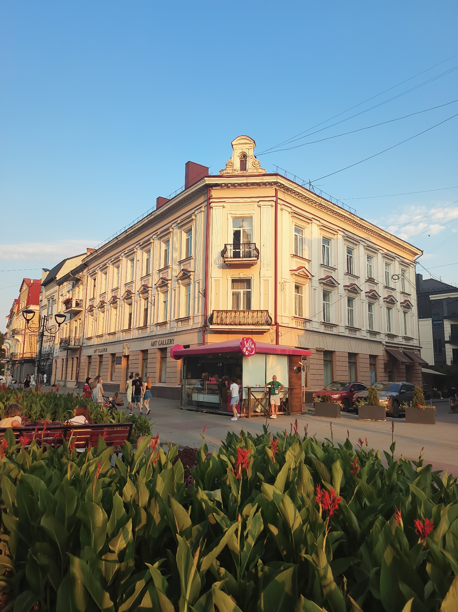 The image depicts a street scene in Ternopil, Ukraine, likely during the late afternoon due to the warm, golden light bathing the buildings. The foreground is dominated by lush green foliage with prominent red flowers, suggesting a park or decorative planting area. A dark wooden bench is visible behind the plants, with several individuals seated on it, facing away from the camera.
Beyond the greenery, a street runs horizontally across the frame, lined with several multi-story buildings. The main building on the right is a light-colored, corner structure with classical architectural details, including columns and ornate window embellishments. A pink and white kiosk with the word "Jolly" is situated in front of this building. Several people are walking on the sidewalk. Two cars are parked on the street in front of the buildings. The sky above is a clear, bright blue. The overall impression is one of a vibrant urban setting with an emphasis on pedestrian activity and civic beautification.