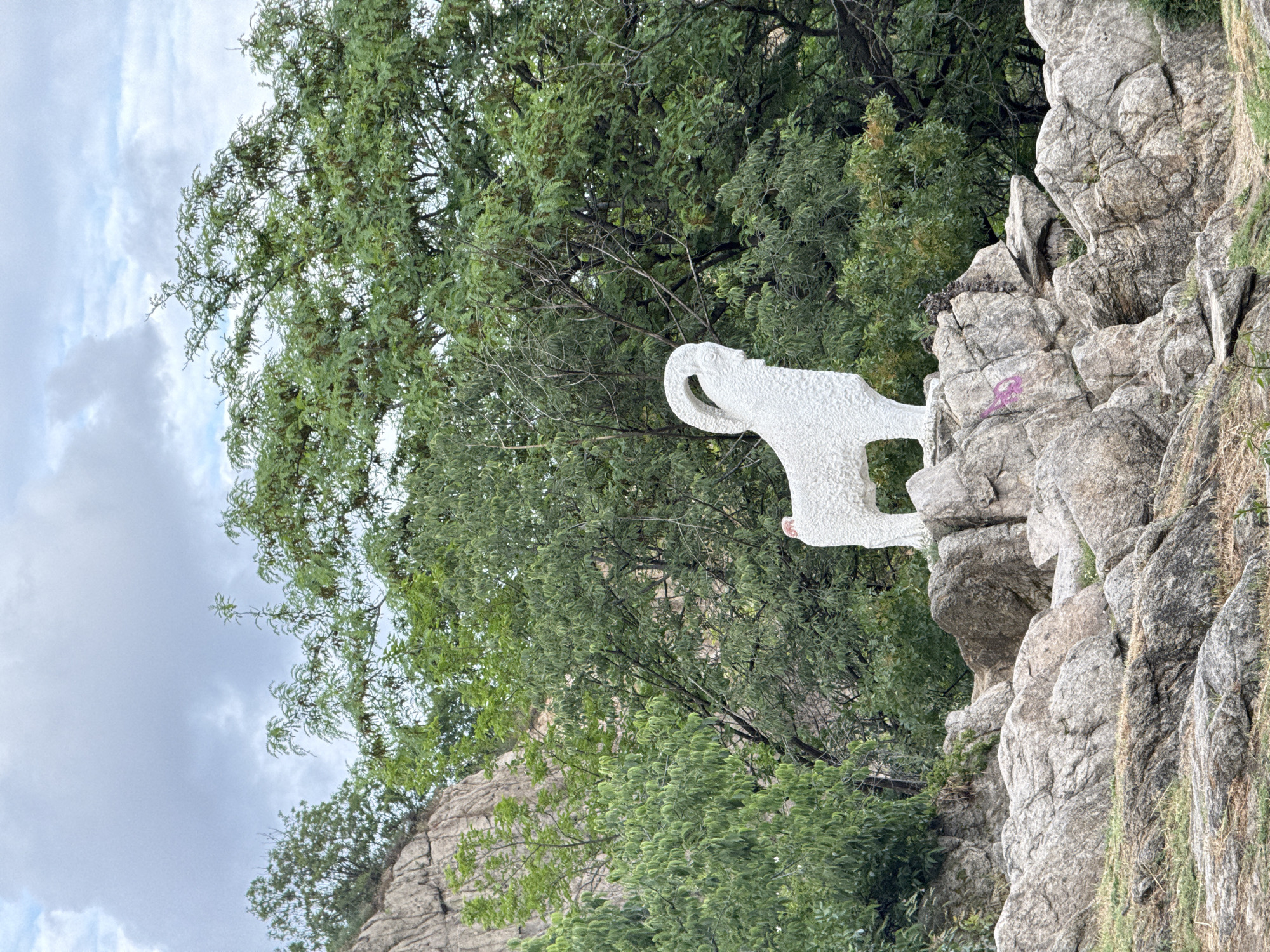 The image shows a white, abstract sculpture of a possible bear on a rocky outcrop. Green trees surround the rocky area. The sky is cloudy. This is the Bear statue on Nebet Tepe in Plovdiv, Bulgaria. There is graffiti on the rocks.