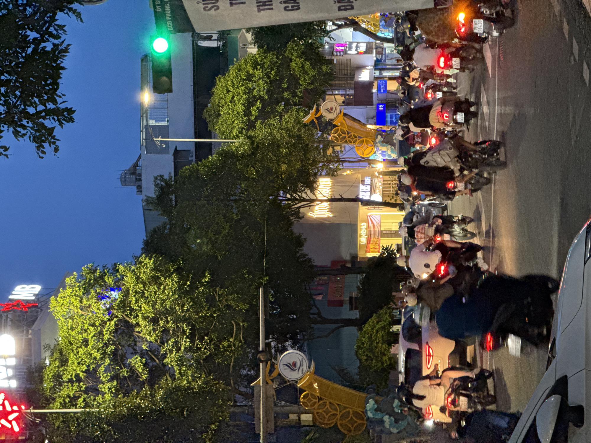 The image depicts a busy street scene in Nha Trang, Vietnam, at dusk. A traffic light is illuminated green, indicating the flow of traffic. Numerous motorbikes are navigating the street, with many riders wearing helmets. The street is lined with trees and buildings, some of which display illuminated signs and advertisements. The overall atmosphere suggests a bustling urban environment during the evening hours.
