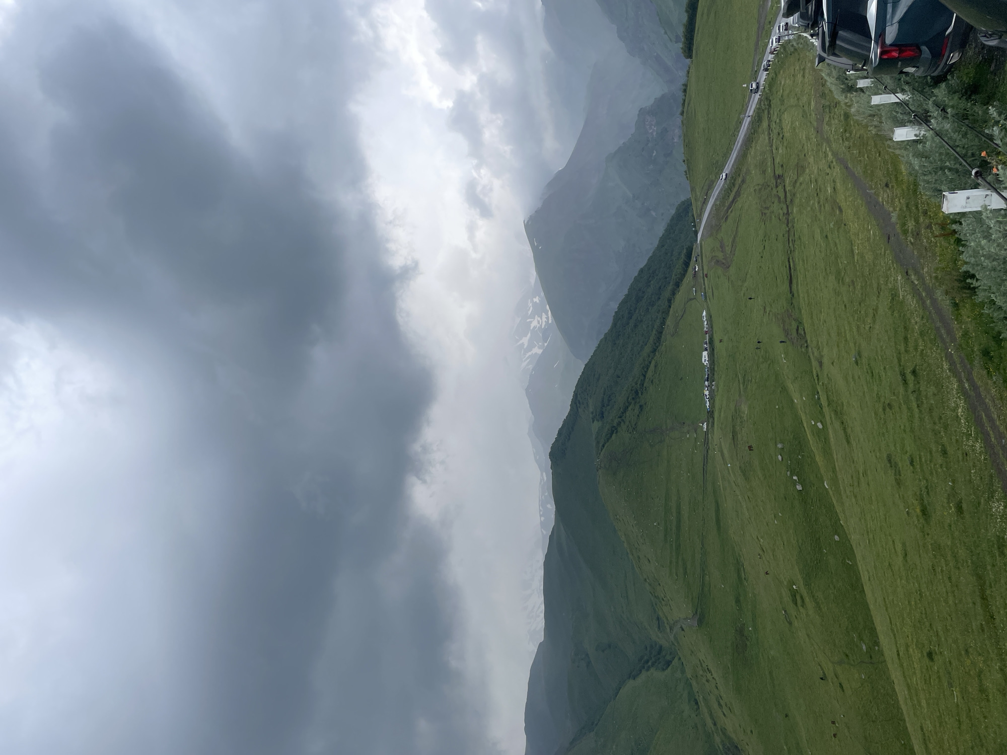 This is an outdoor, high-angle, full shot of a cloudy, mountainous landscape in Stepantsminda, Georgia. The foreground features several parked vehicles, including the rear of a dark SUV, along a roadside barrier. Beyond the barrier, a green, grassy slope descends, dotted with a few scattered stones and what appear to be animals, possibly horses, grazing. A dirt path winds its way down the slope. In the midground, a paved road curves into the distance, with a line of cars and a few people visible along its edge. More grassy hills and mountains stretch into the background, some with patches of snow on their peaks. The sky is filled with dramatic, dark grey storm clouds, obscuring much of the sky and creating a moody atmosphere. The overall scene depicts a natural, rugged environment under overcast, possibly stormy, weather conditions.