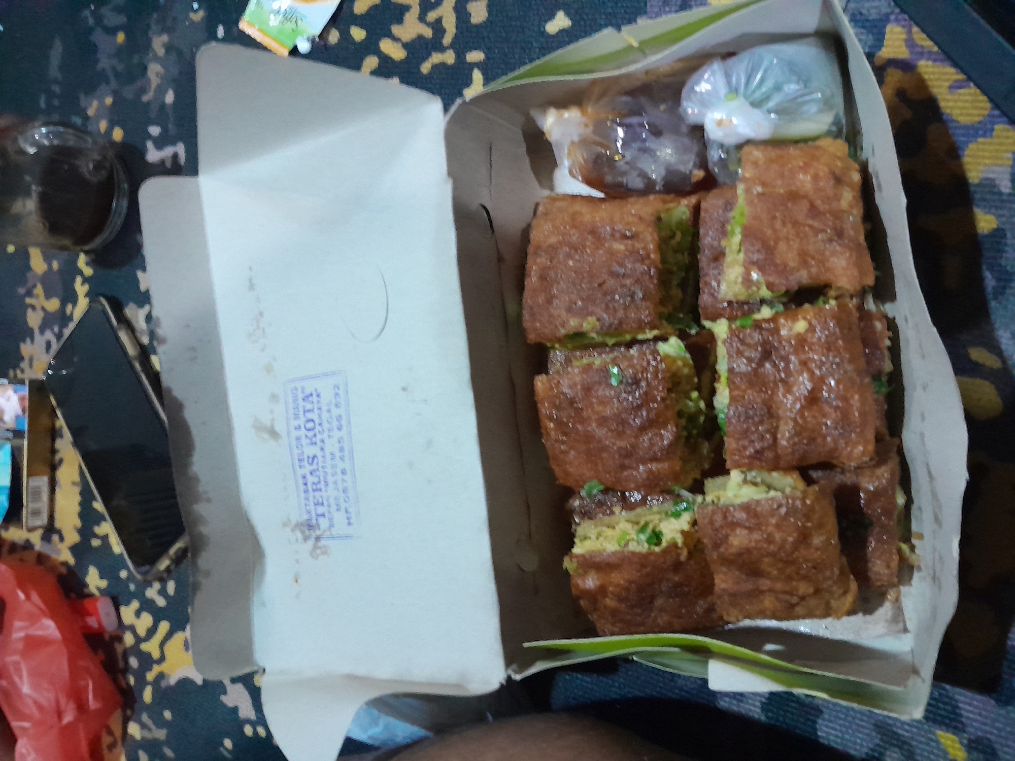 An overhead medium shot shows a opened white cardboard box containing several pieces of murtabak, a savory Indian-style pancake filled with meat, on a patterned surface. The murtabak are cut into rectangular portions and have a golden-brown, slightly crispy exterior. Some green scallions are visible within the filling of one piece. To the right of the murtabak, a small plastic bag contains a dark sauce, and another small plastic bag holds a white, round object, possibly a pickled vegetable. The inside of the box lid has a blue and white stamp with text in Indonesian, reading "MARTABAK TELOR & MANIS TERAS KOTA" and "JL. JASEN - TEGAL". In the background, a red plastic bag, a smartphone, and a glass of dark liquid are visible. The scene appears to be set indoors or on an outdoor table.