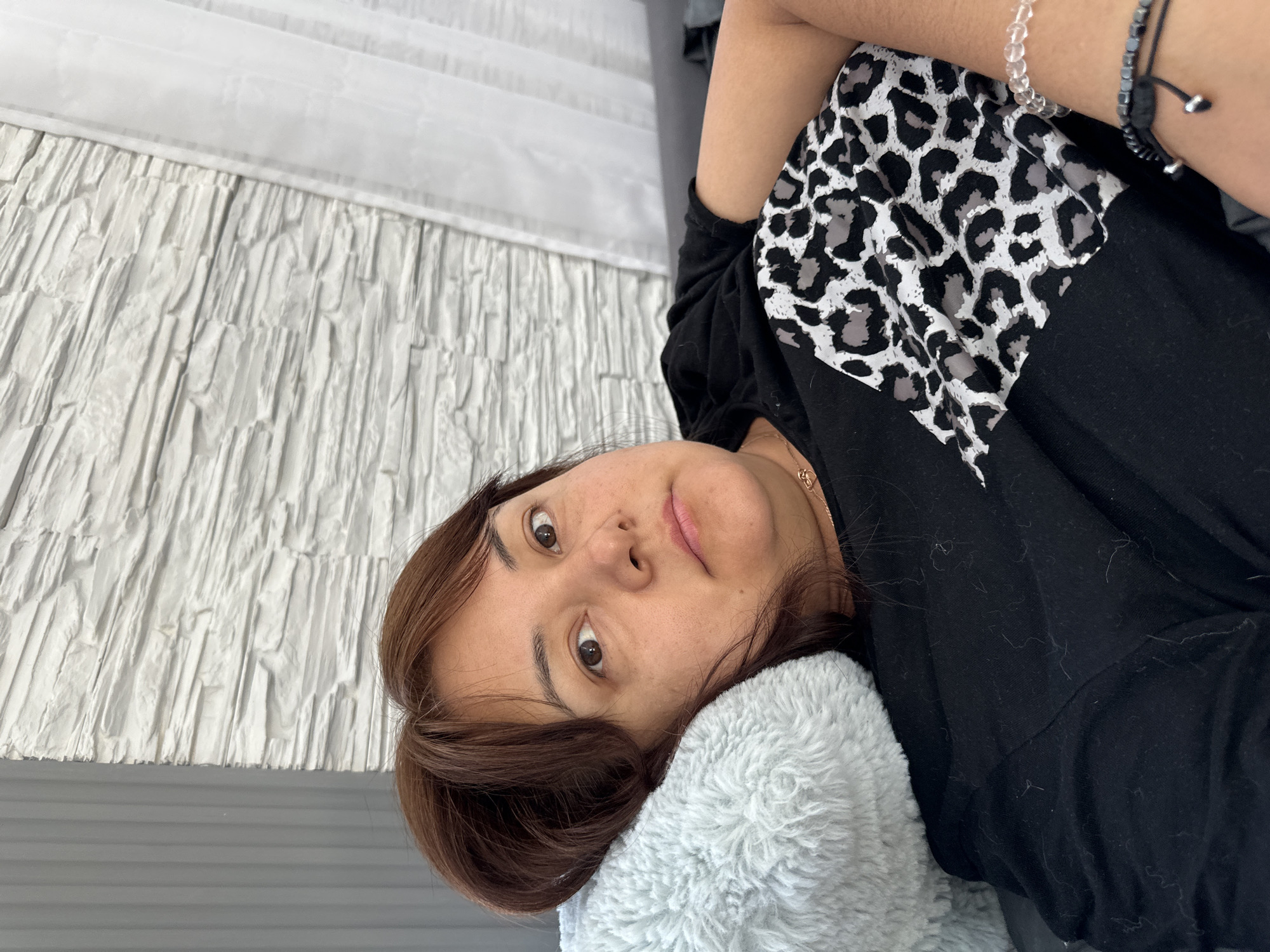 A woman lies on a light grey, fluffy pillow against a white headboard. She is wearing a black t-shirt with a black and white animal print accent on the upper left. She has brown hair and brown eyes. Two bracelets are visible on her raised left wrist. The location is a bedroom in an unknown city.
