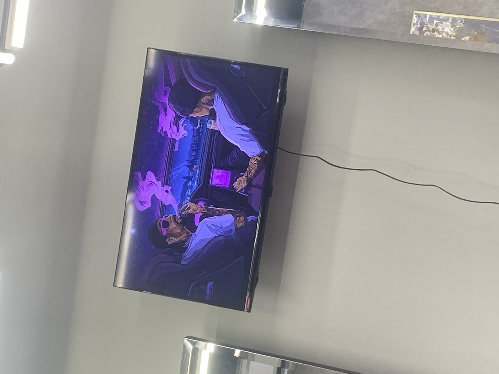 A television is mounted on a wall, displaying a cartoon illustration of two people in an urban environment. One figure, seated, wears sunglasses and is smoking. The other figure stands behind, leaning in. The image appears to be indoors, with a white wall as the background. A power cord extends from the television. The setting is a room in Kuta, Indonesia.