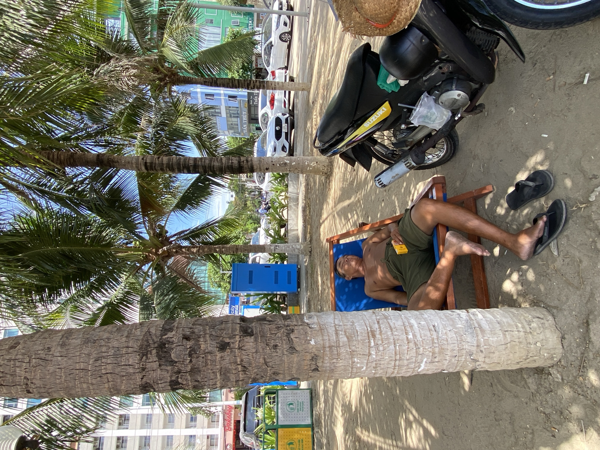 A shirtless man in green shorts is sleeping on a blue beach chair on a sandy beach in Nha Trang, Vietnam. He is leaning back, with his head resting on the chair. A yellow cigarette pack is on his chest. A large palm tree trunk is to the left of the frame, providing shade. To the right of the frame, a black motorcycle is parked on the sand, with a straw hat on its seat. In the background, there are several buildings, palm trees, and parked cars. The sky is blue and clear, indicating a sunny day.