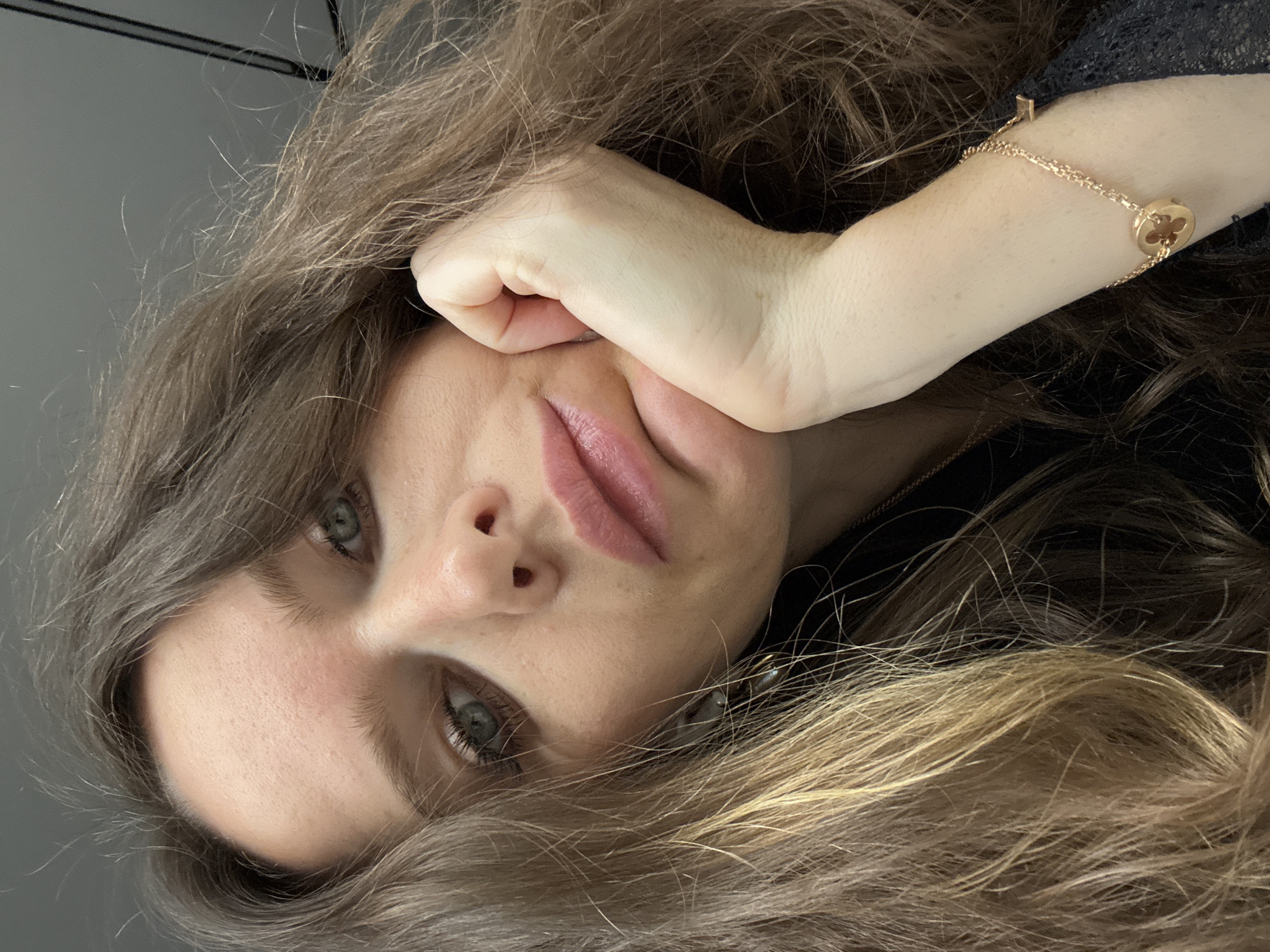 Close-up portrait of a Caucasian woman with long brown hair, partially blonde towards the ends, resting her face on her hand. She has fair skin, prominent cheekbones, and light-colored eyes. She is wearing a delicate gold bracelet with a circular pendant. The background appears to be a neutral gray color. There is no information to suggest this photo was taken in Moscow, Russia.