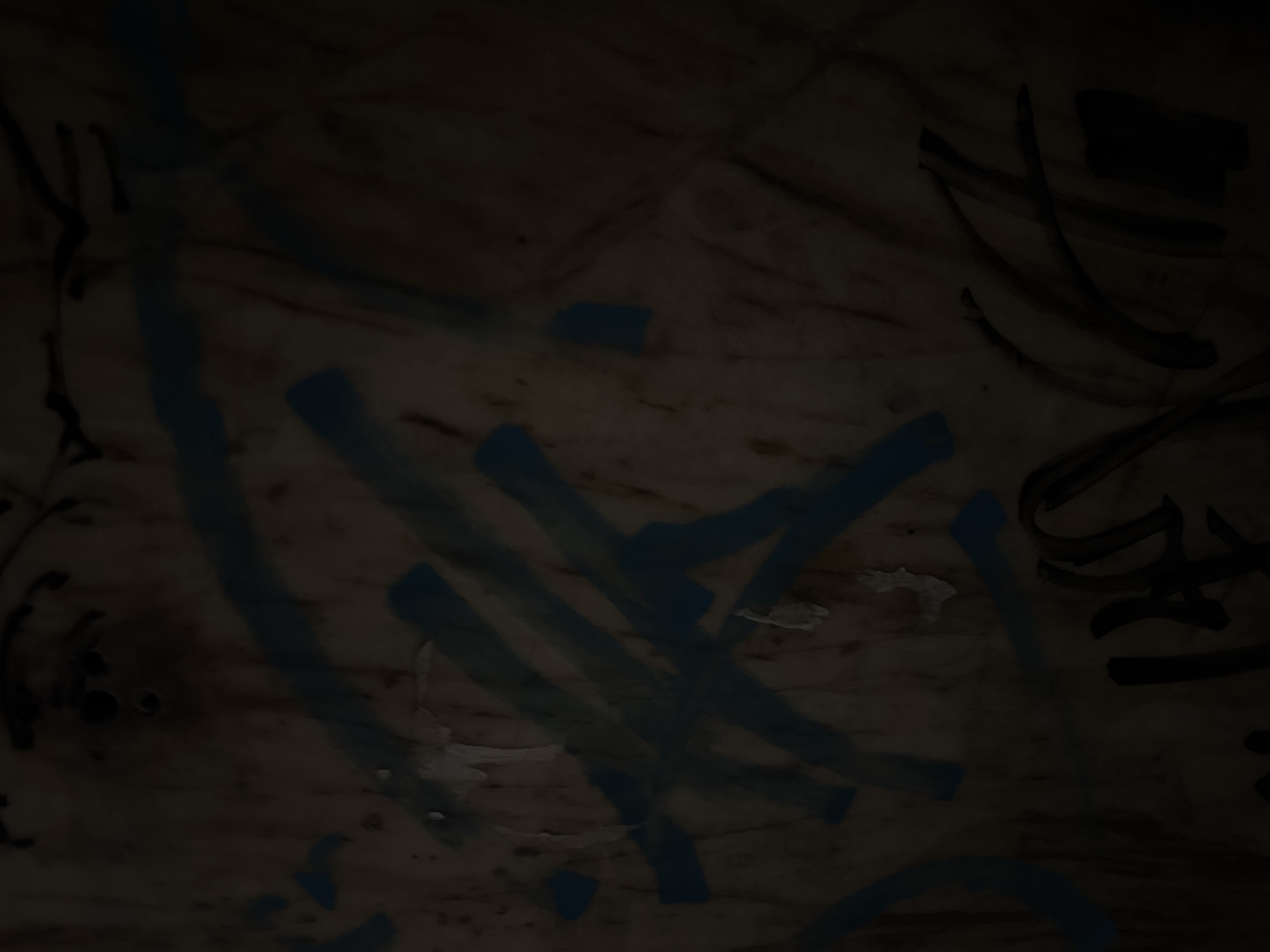 The image displays graffiti on a textured surface, likely a wall or wooden panel, in the Vracar Urban Municipality, Serbia. The graffiti consists of blue spray-painted lines and scribbles, superimposed on darker, possibly black, markings. The surface appears worn and weathered, with some areas showing peeling or damaged material. The lighting is dim, casting shadows and obscuring some details of the graffiti and the underlying surface. There are no visible people or other discernible subjects in the frame.
