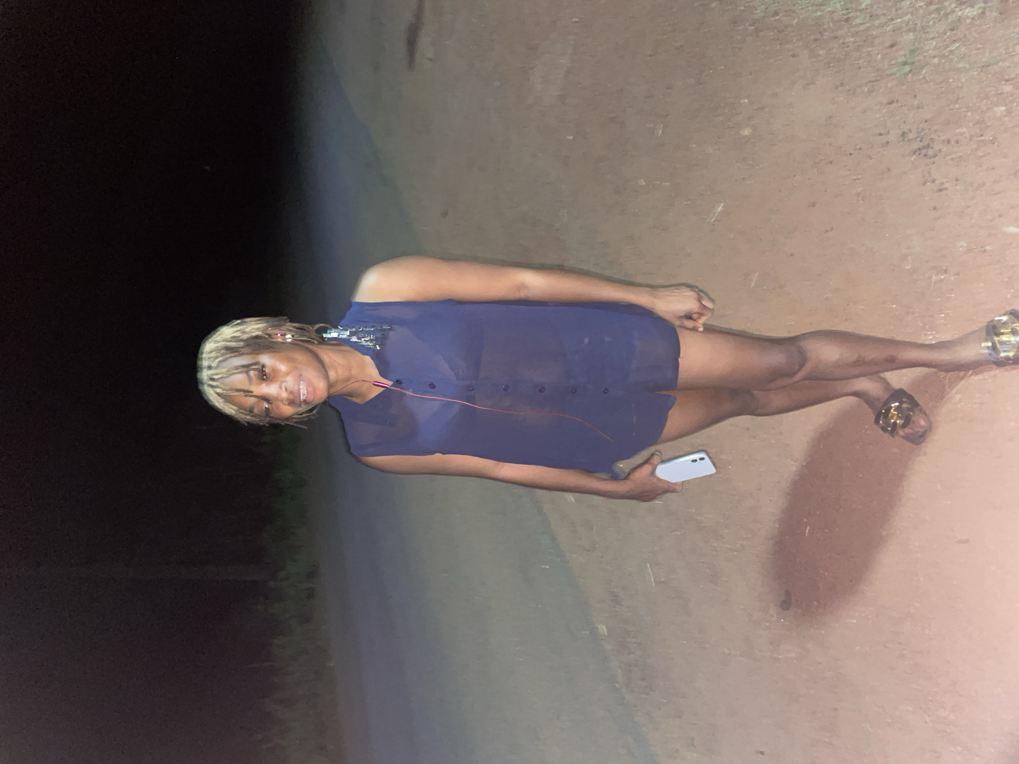 The image shows a woman standing on a dirt road at night. The location is possibly near Ogbe, Nigeria, based on available information. The woman is wearing a dark, translucent blouse and appears to be holding a phone. The surrounding environment consists of dark areas with potential vegetation or trees in the background, and a lighter-colored dirt surface under her feet. The overall lighting suggests the use of a camera flash or dim ambient light.