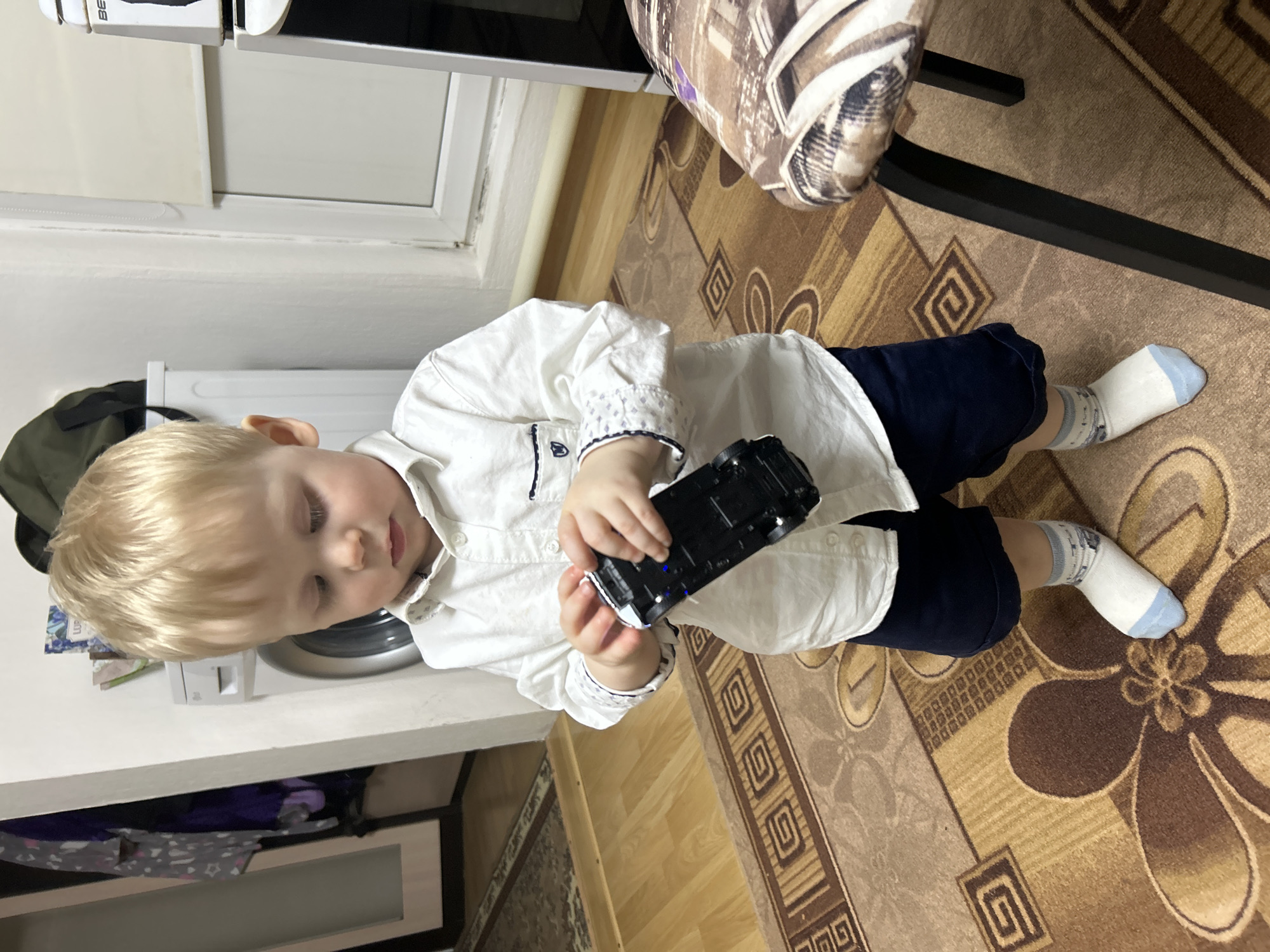 An indoor scene in a residential setting in the city of Фоки, Russia, depicts a young, blonde-haired child standing centrally, looking downwards. The child is wearing a white collared long-sleeved shirt with navy trim and navy shorts, along with white socks featuring light blue toes and heels, and a gray patterned ankle band. The child holds a black toy car with white/silver details and visible blue accents on its underside, examining it with both hands.
The floor is covered with a patterned carpet in shades of brown and tan, displaying geometric and floral designs. To the left, a light-colored doorframe reveals a wooden floor beyond, with draped clothing visible. Behind the child and to the left, a white washing machine is partially visible, with an olive-green bag and papers resting on a surface above it. To the right, a white-framed window with a drawn light-colored roller blind is present, next to a portion of a tall white appliance, possibly a refrigerator. In the immediate foreground to the right, a black metal chair leg and the edge of a chair seat with a brown and white patterned fabric cover are visible. The room is illuminated by artificial lighting.