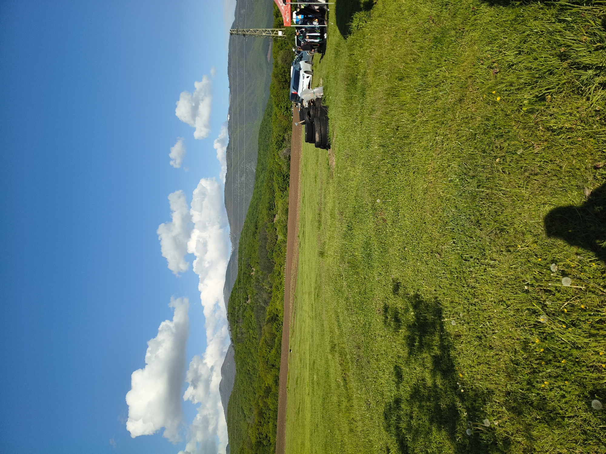The scene is in Topolovo, Bulgaria. A grassy field, with a patch of dirt, runs alongside a wooded hill. Mountains are in the background. The sky is blue with puffy white clouds. There are people and vehicles parked near the edge of the field. Discarded tires are lying on the ground. There is a power line structure visible.
