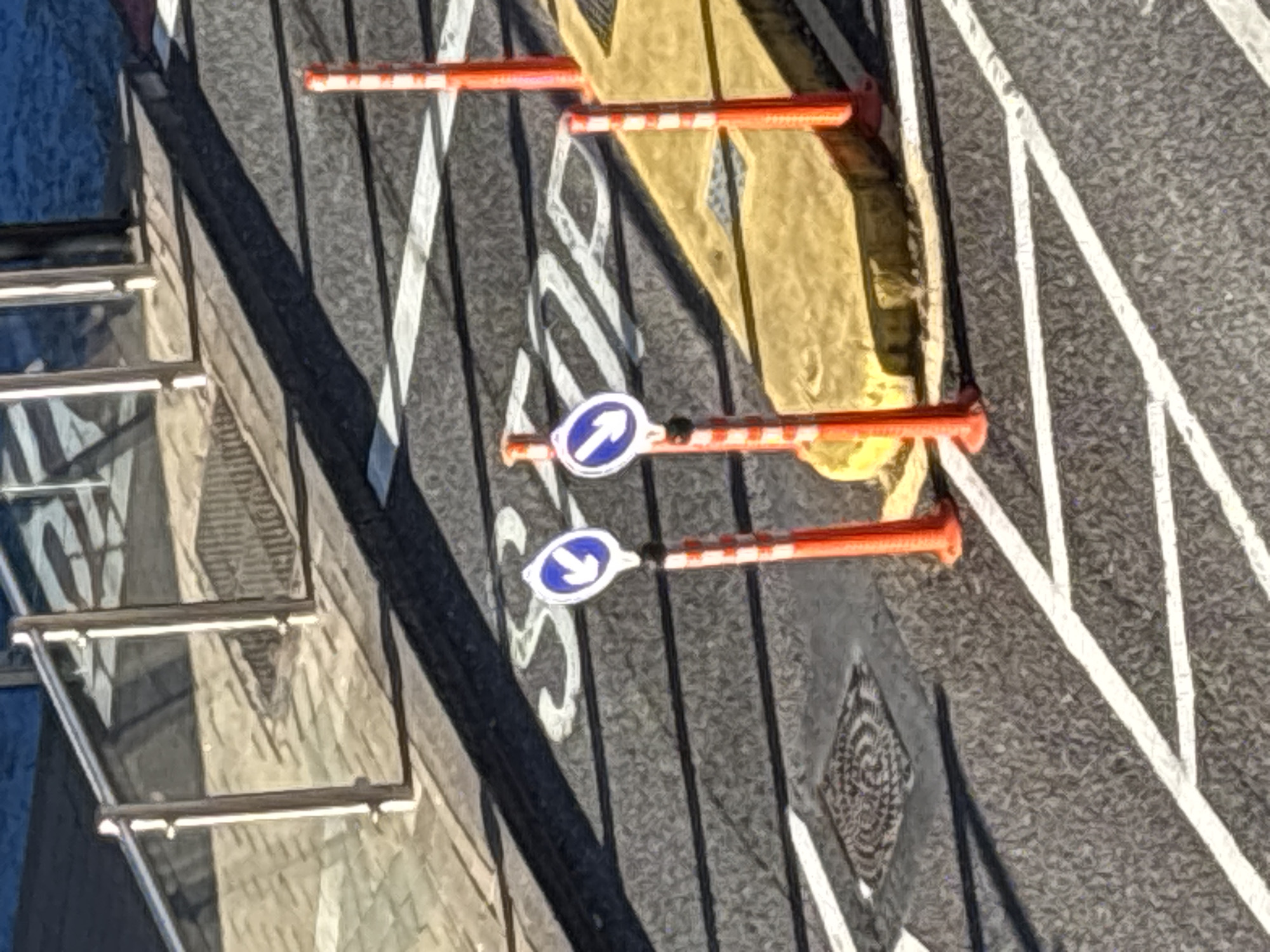 The image shows a road surface in Cork, Ireland, with traffic markings and street furniture. Two white arrows with blue circular backgrounds, indicating direction, are mounted on orange and white striped posts. These posts are positioned on a yellow concrete island which is adjacent to a pedestrian crossing. Further down the road, there are white line markings, some forming a pedestrian crossing, and a manhole cover. In the background, a metal railing and a shadow suggest the presence of a structure, possibly a building or a bridge. The scene is outdoors, on a paved road surface, under daylight conditions.