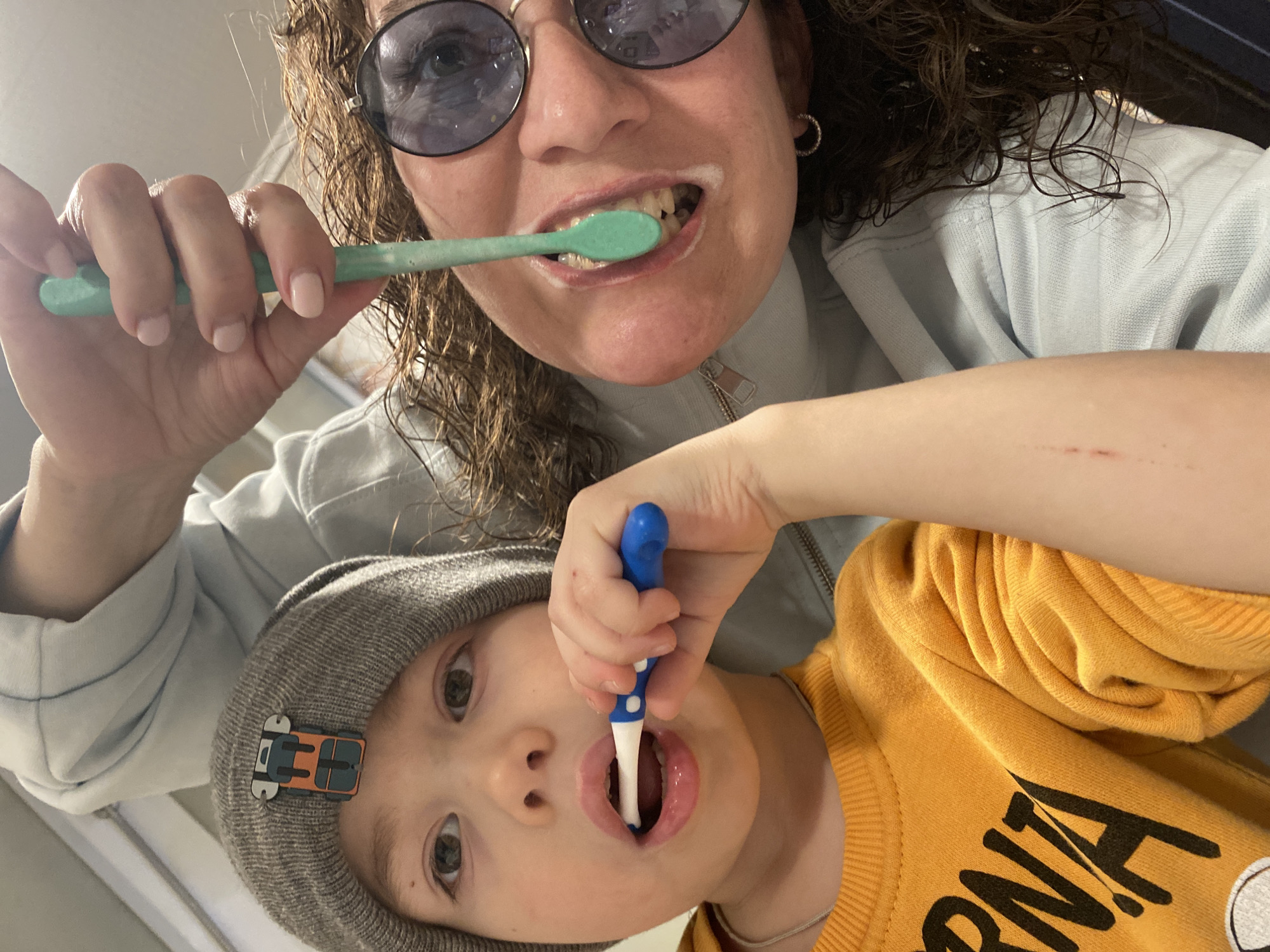 A woman and a child are brushing their teeth. The woman is wearing round, dark-tinted glasses, curly hair, a light blue jacket and hoop earrings. The child is wearing a gray beanie with a rectangular clip-on attachment, and an orange sweatshirt with black lettering. Both hold their toothbrushes up close to the camera, with toothpaste visible on the woman's teeth. The location is unknown and there is no evidence suggesting it is in Ольховка, Russia.