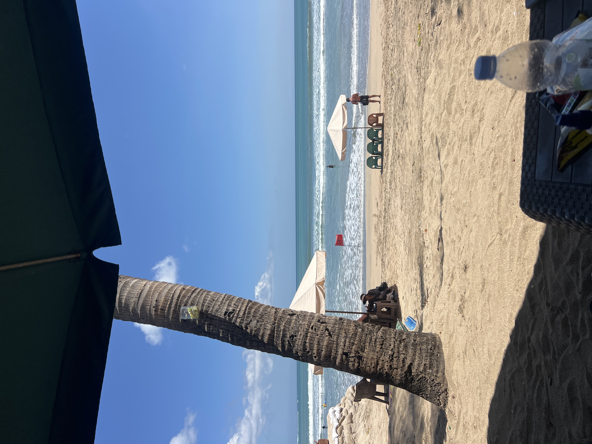 The image depicts a beach scene in Kuta, Indonesia, on a sunny day with a clear blue sky and white clouds. In the foreground, a dark green umbrella casts a shadow over the sandy beach. A large palm tree trunk, with a plastic bag containing what appears to be fruit tied to it, stands to the left. Further back, a beige umbrella provides shade for two people sitting on chairs, with a red flag planted nearby in the sand. Beyond them, the ocean stretches to the horizon, with gentle waves breaking on the shore. In the middle ground, three green plastic chairs and one orange plastic chair are arranged in front of another beige umbrella, where a man in black shorts stands looking out at the water. A person is visible in the water in the distance. The sand is light brown and marked with footprints. In the bottom right corner, a plastic water bottle and some snack bags are placed on a dark woven surface, suggesting a relaxed atmosphere.
