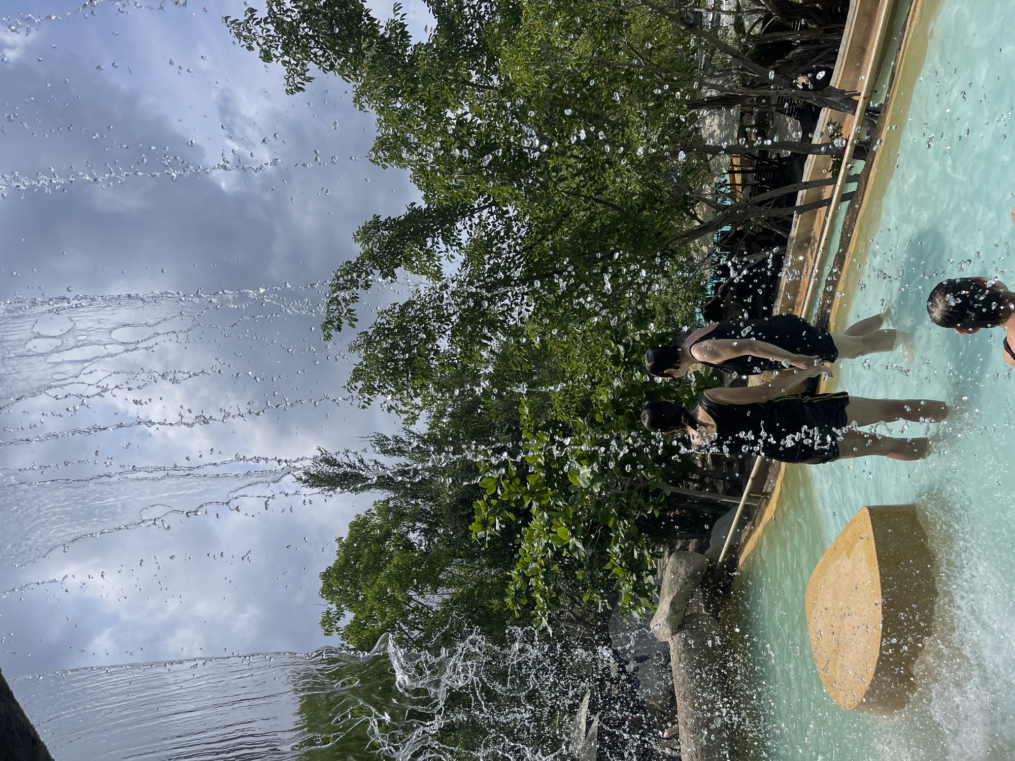 Two individuals are standing in a pool with light blue water, possibly at a water park or recreational area. The pool is enclosed by a curved, tiled edge. The people are positioned near what appears to be a large, light-colored rock formation or structure within the pool. In the foreground, a curtain of water is falling, creating a blurred effect due to the motion. In the background, there are trees, foliage, and what seems to be a railing or fence. The sky above is partly cloudy. The location is identifiable as Tây Nha Trang, Vietnam due to the specific foliage and landscaping in the image, but confirmation would require more information. The image does not clearly depict the people’s actions or interactions in great detail.