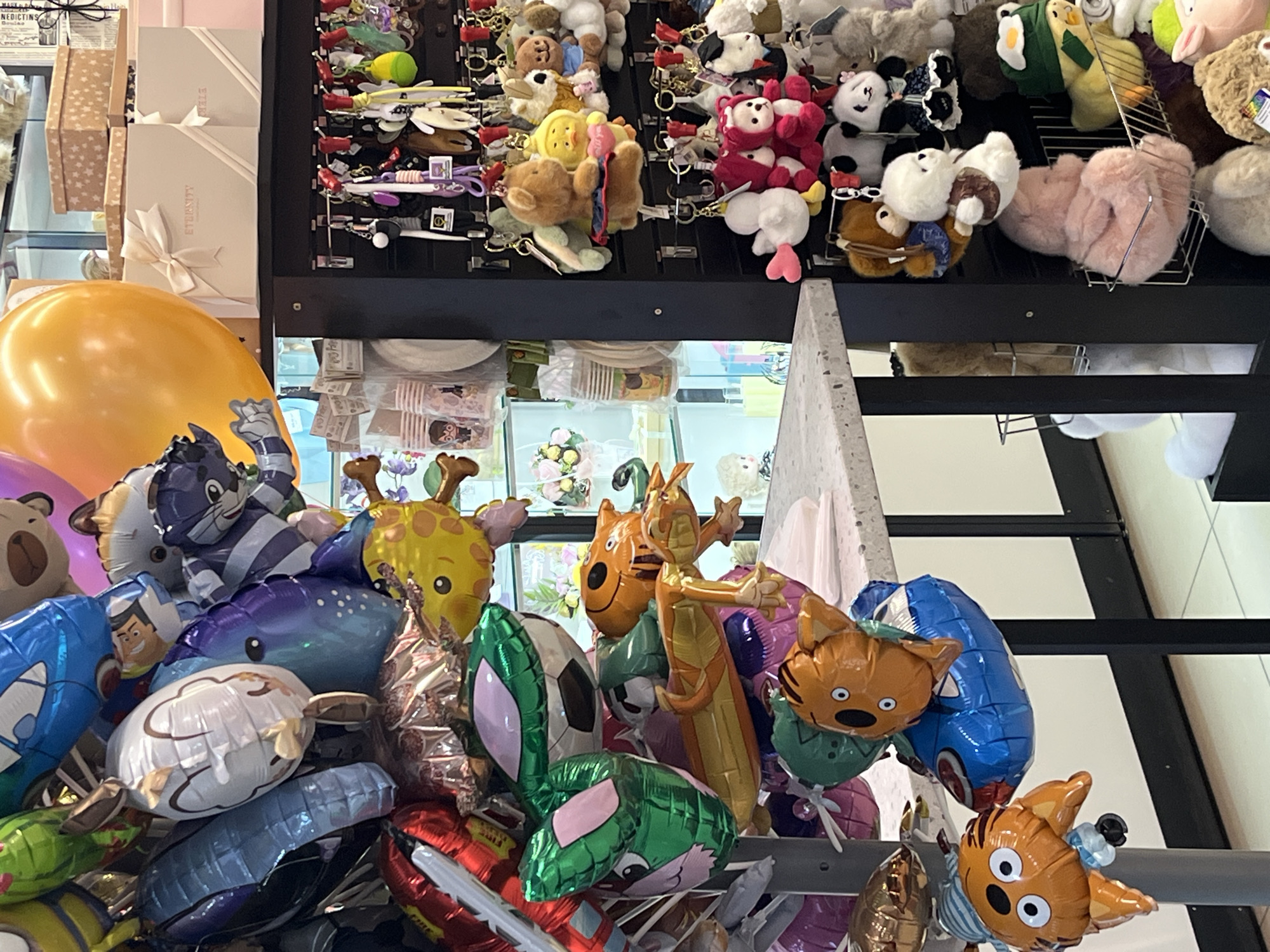 The image shows a display of balloons and stuffed animals. The balloons are various shapes and colors, including characters like a wolf, giraffe, and tiger, and some are in the shape of vehicles and a soccer ball. The stuffed animals above the balloons are small and appear to be keychains. Boxes are visible in the background, as well as a window. There is no indication of a specific location in Izhevsk, Russia, within the image.